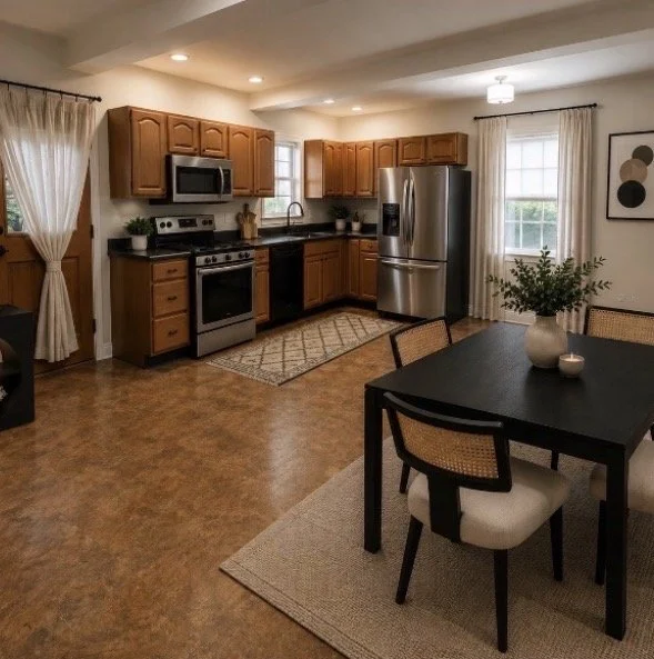 Kitchen with wooden cabinets, stainless steel refrigerator, microwave, oven, and dark countertop. Adjacent dining area with black table, four chairs with beige cushions, a white vase with green plant, and framed artwork on the wall. Light-colored curtains and hardwood flooring.