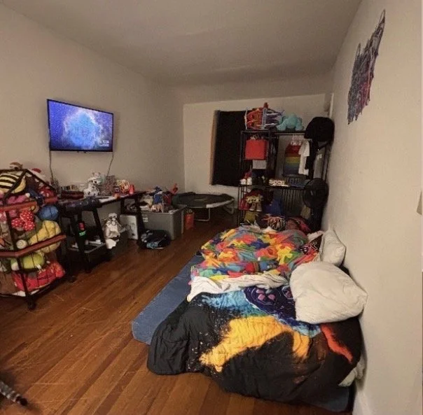 A cluttered bedroom with a bed covered in colorful blankets on the right, a TV mounted on the wall on the left, and toys and items scattered on a shelving unit and around the room.