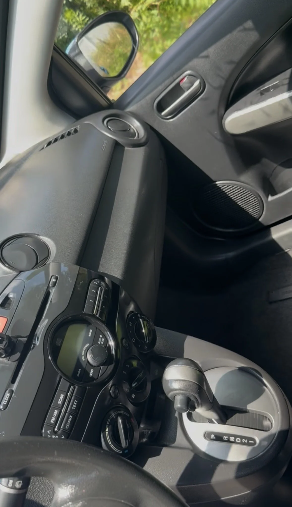 Inside view of a car dashboard with various controls, a gear shifter showing 'R D 2 1', and a side mirror reflecting greenery outside.