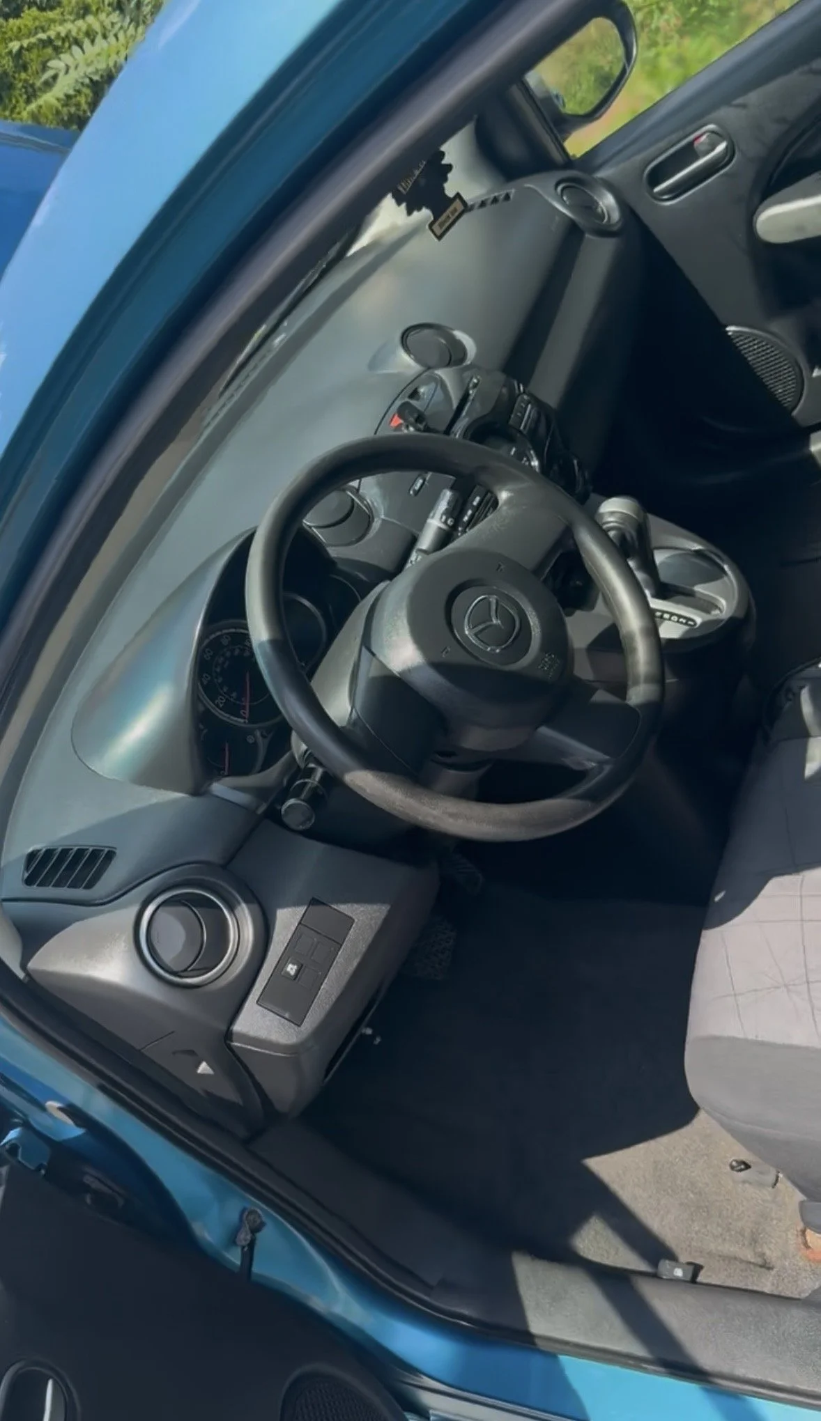 Interior of a small blue car showing the steering wheel, dashboard, control buttons, and a view of the side mirror.