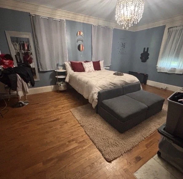A bedroom with a blue accent wall, a bed with red and white pillows, a chandelier ceiling light, a gray ottoman at the foot of the bed, and a wooden floor with a beige rug.