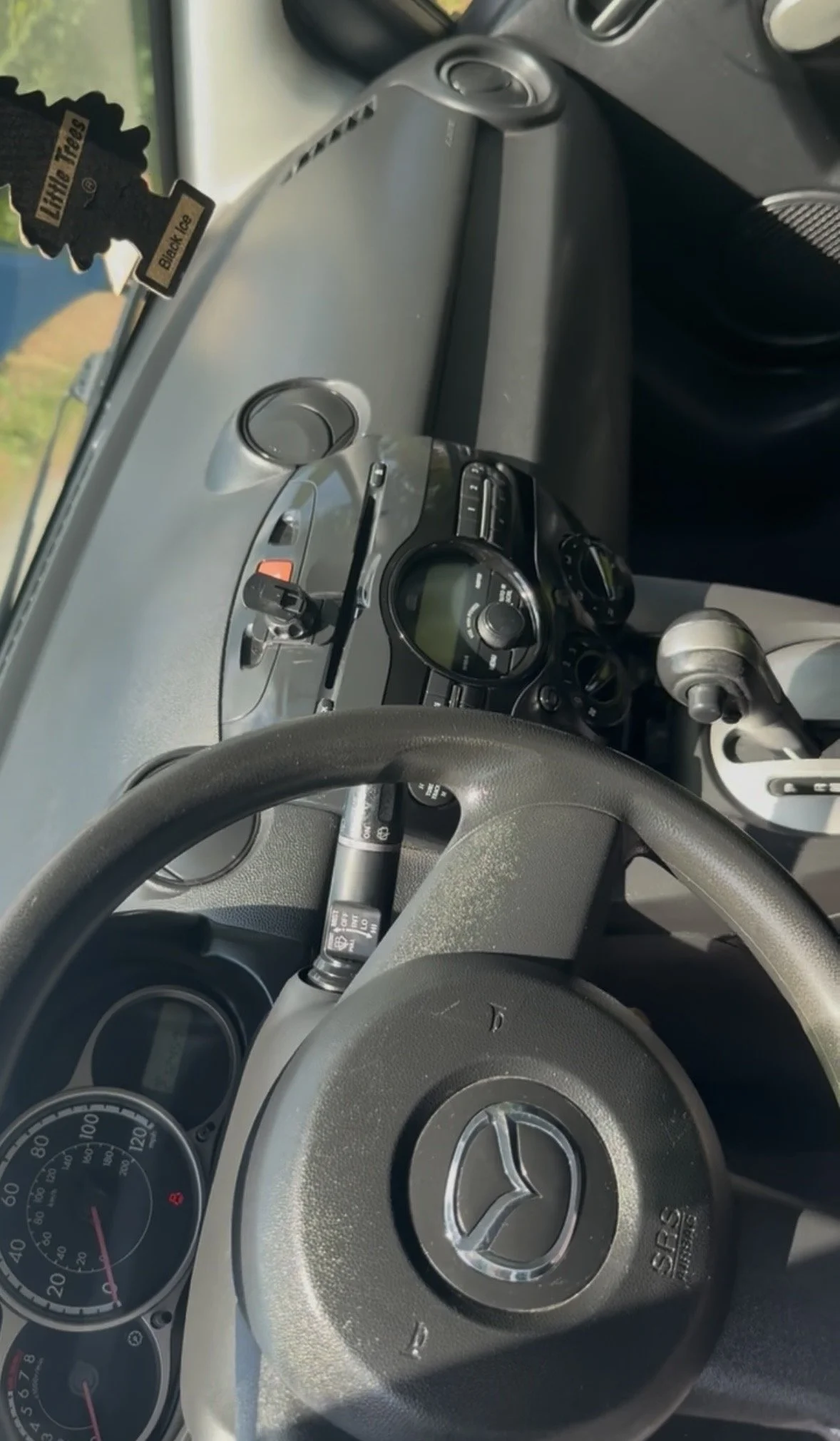 The image shows the interior dashboard of a Mazda vehicle, including the steering wheel with Mazda logo, speedometer, and air conditioning vents.
