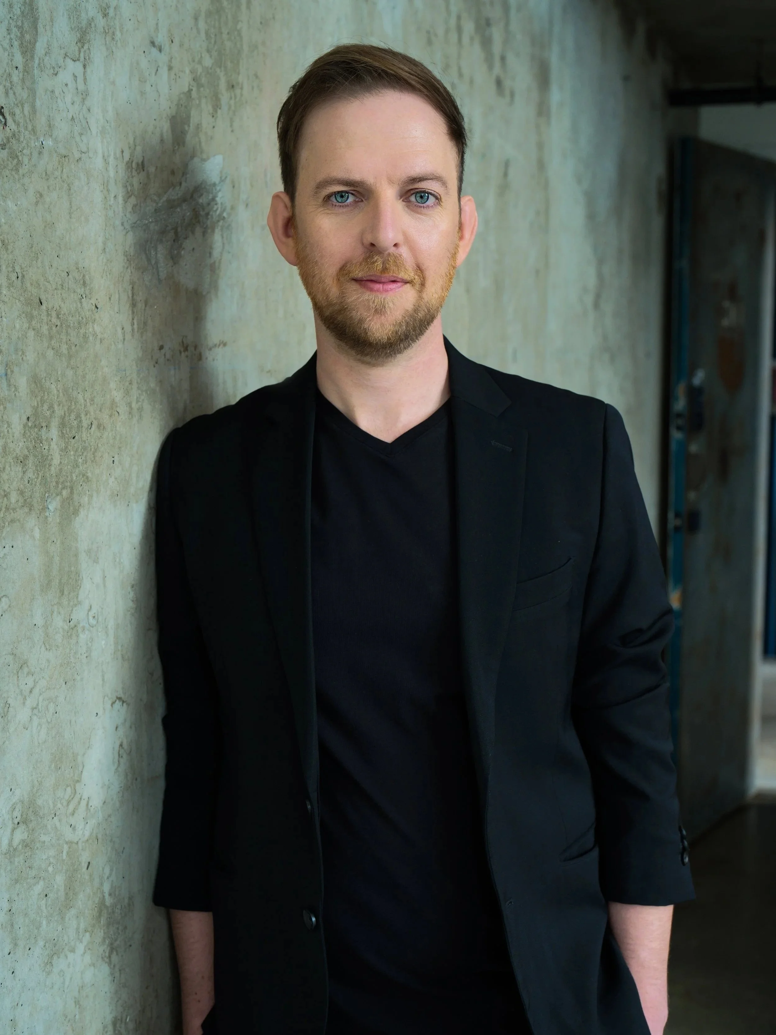 A man with light skin, blue eyes, and brown hair, wearing a black shirt and black blazer, leaning against a weathered concrete wall.