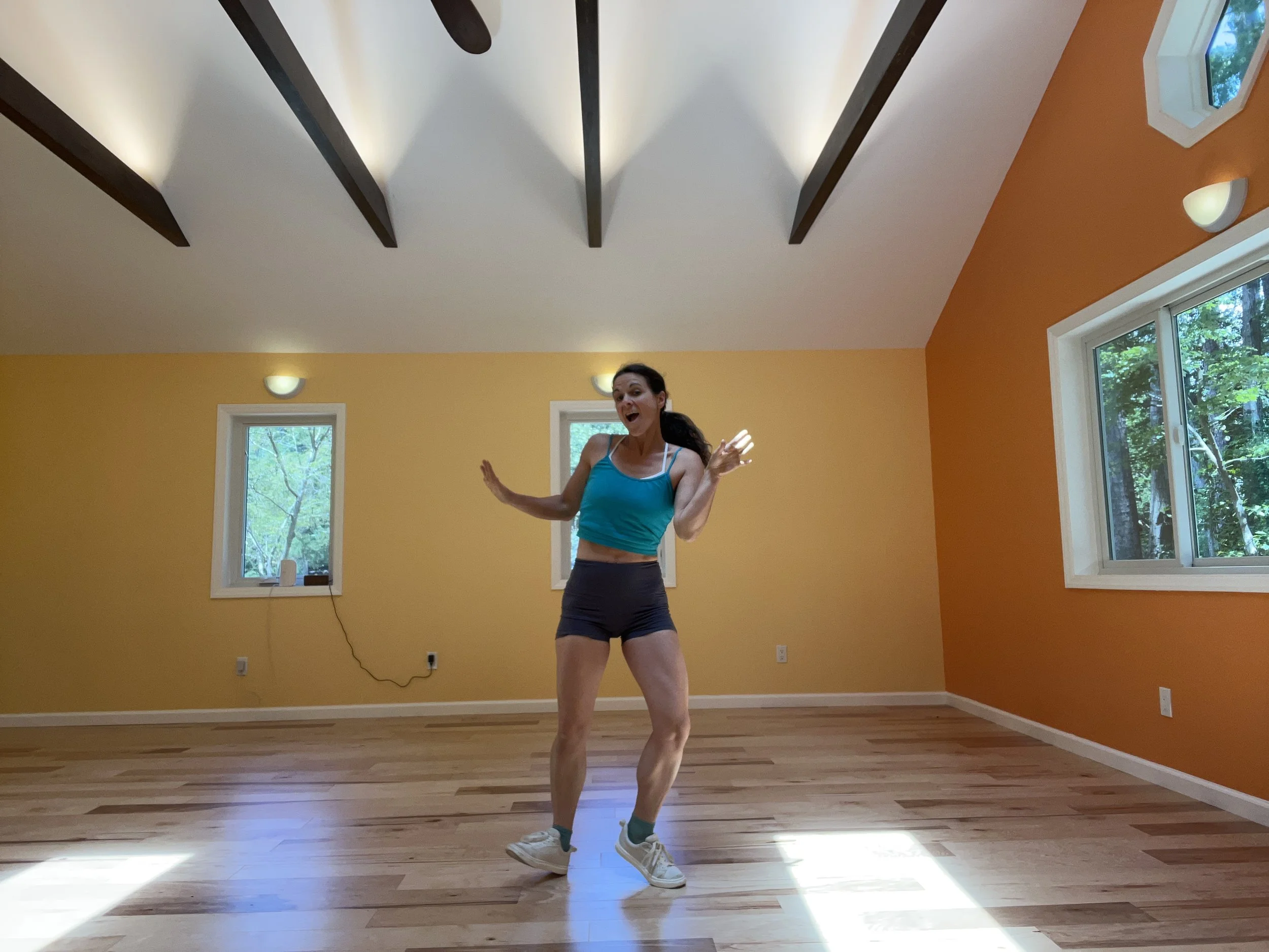 Woman dancing or exercising in a brightly lit room with wooden floors and orange walls, with large windows showing greenery outside.