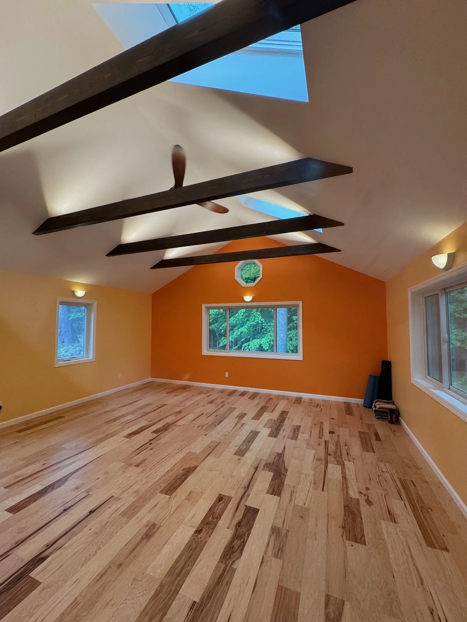 Empty room with light wood flooring, yellow and orange walls, large windows, vaulted ceiling with wooden beams, and a ceiling fan.