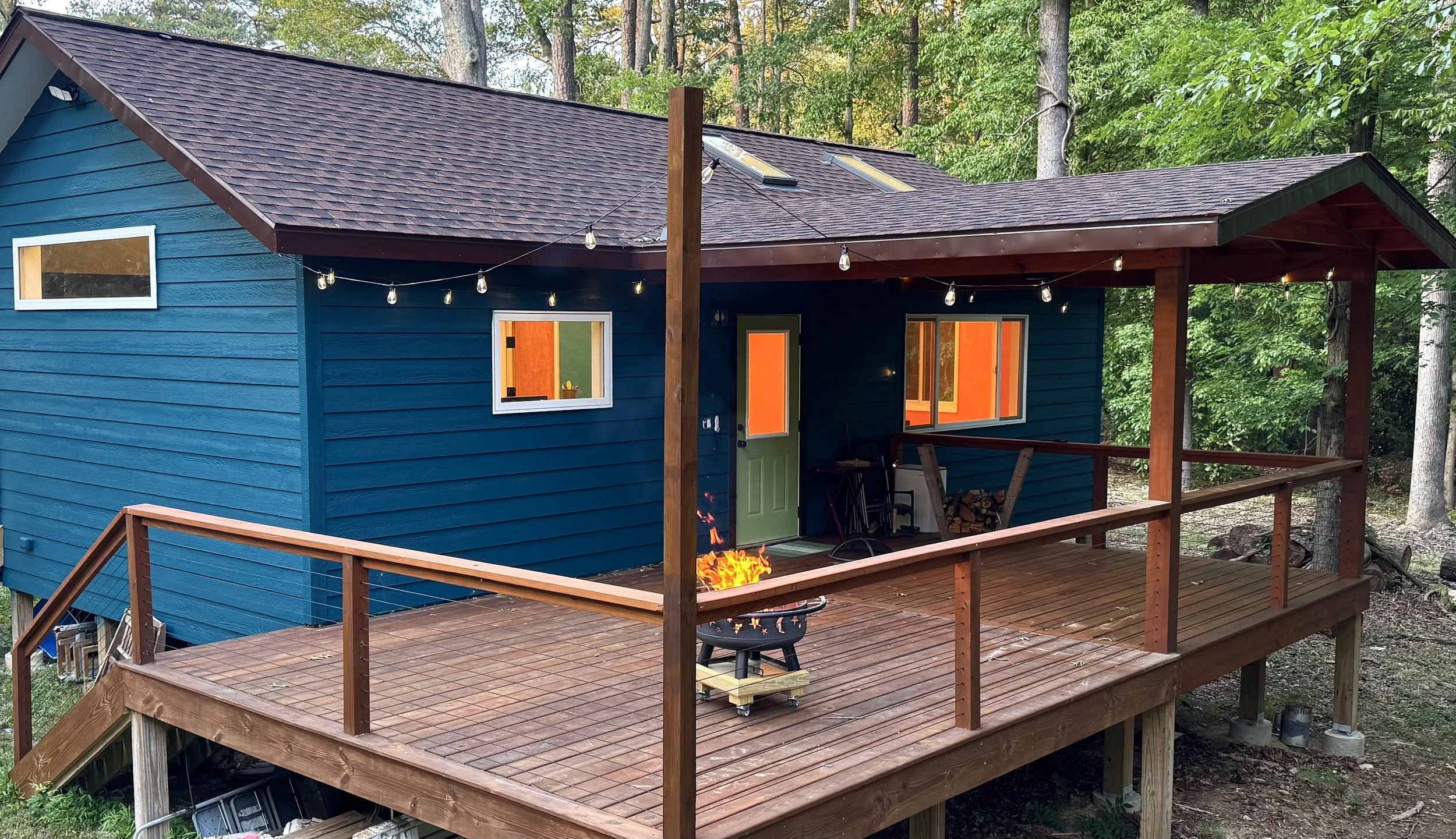 A wooden deck attached to a blue house with a green door, surrounded by trees, and string lights hanging overhead. There's a fire pit with a small fire burning on the deck.