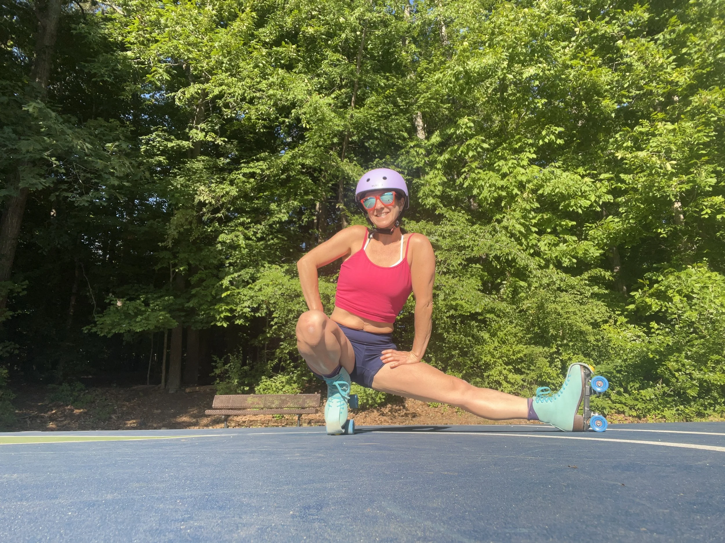 Woman wearing sunglasses, helmet, and pink tank top roller skating on a handstand pose at a park with trees in the background.