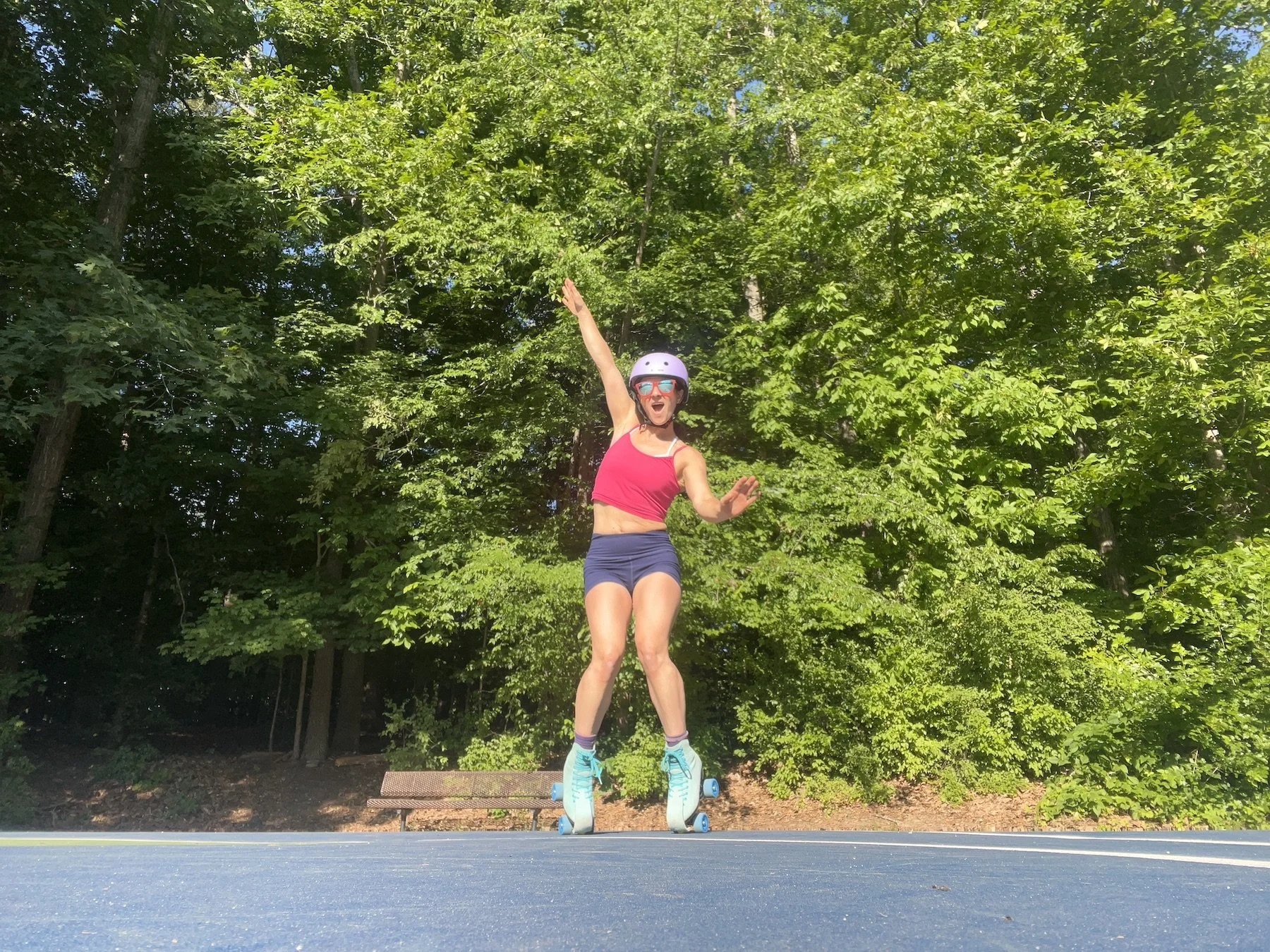 A woman in a pink sports bra, navy shorts, and roller skates balances on a skateboard outdoors surrounded by green trees. She is wearing a helmet and sunglasses, raising one arm and smiling in excitement.