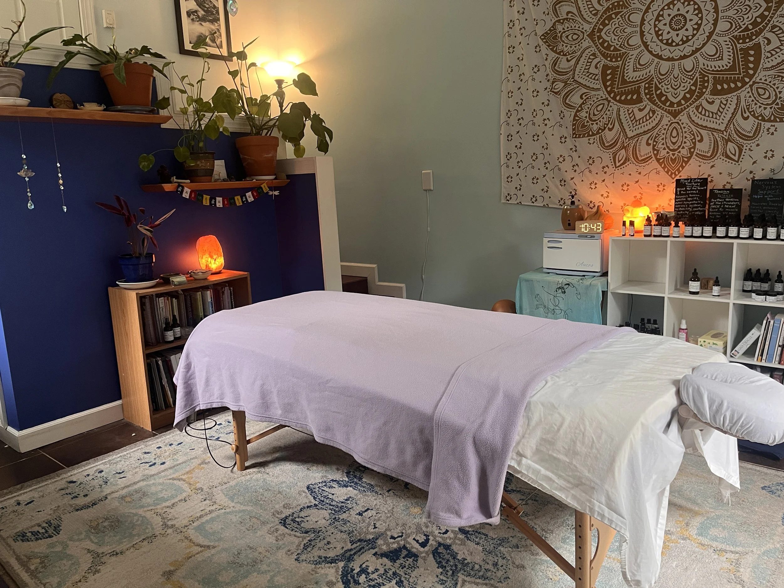 A cozy room with a massage table covered with a lavender sheet, a white blanket, and a pillow. The wall behind has a large mandala tapestry on the right, and a yellow lamp, small wooden figures, and bottles of essential oils are on a white shelf. There are potted plants on wall shelves, framed photos, and decorative hanging crystals on the left. The floor has a patterned rug.