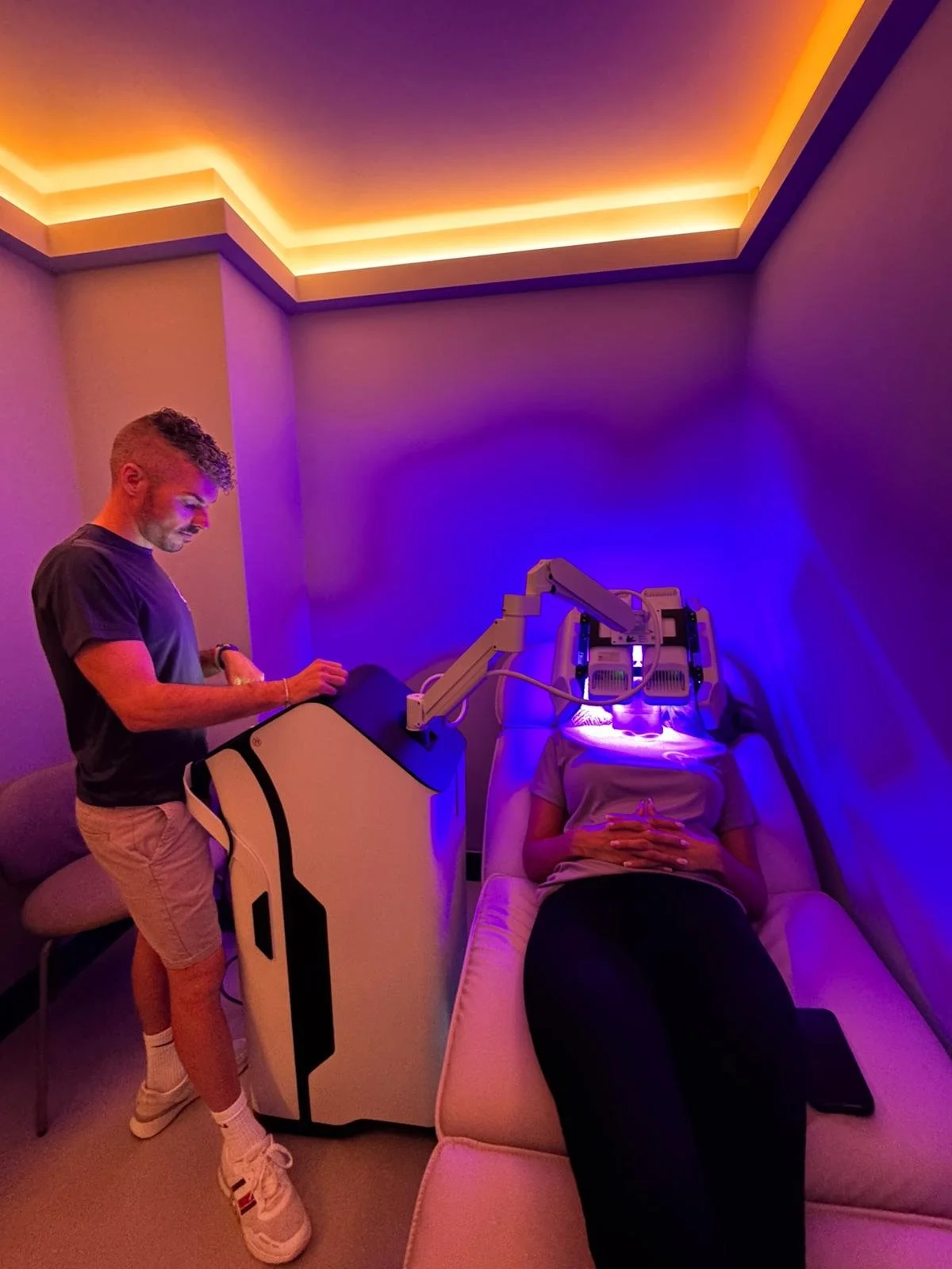 A person lying on a treatment bed with a medical device emitting purple light over their face, while a technician operates the device in a room with colorful lighting.