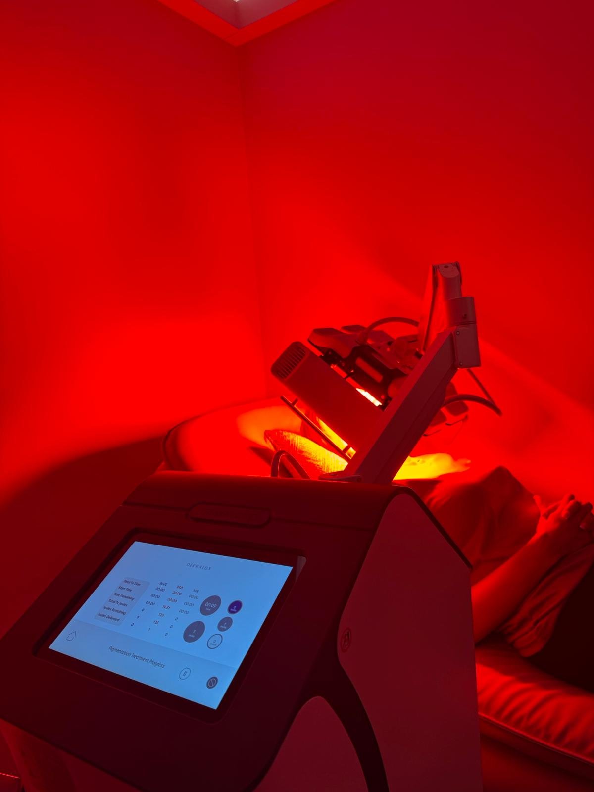 A person lying on a bed receiving a red light therapy treatment with a medical device, with a monitor displaying settings in the foreground.