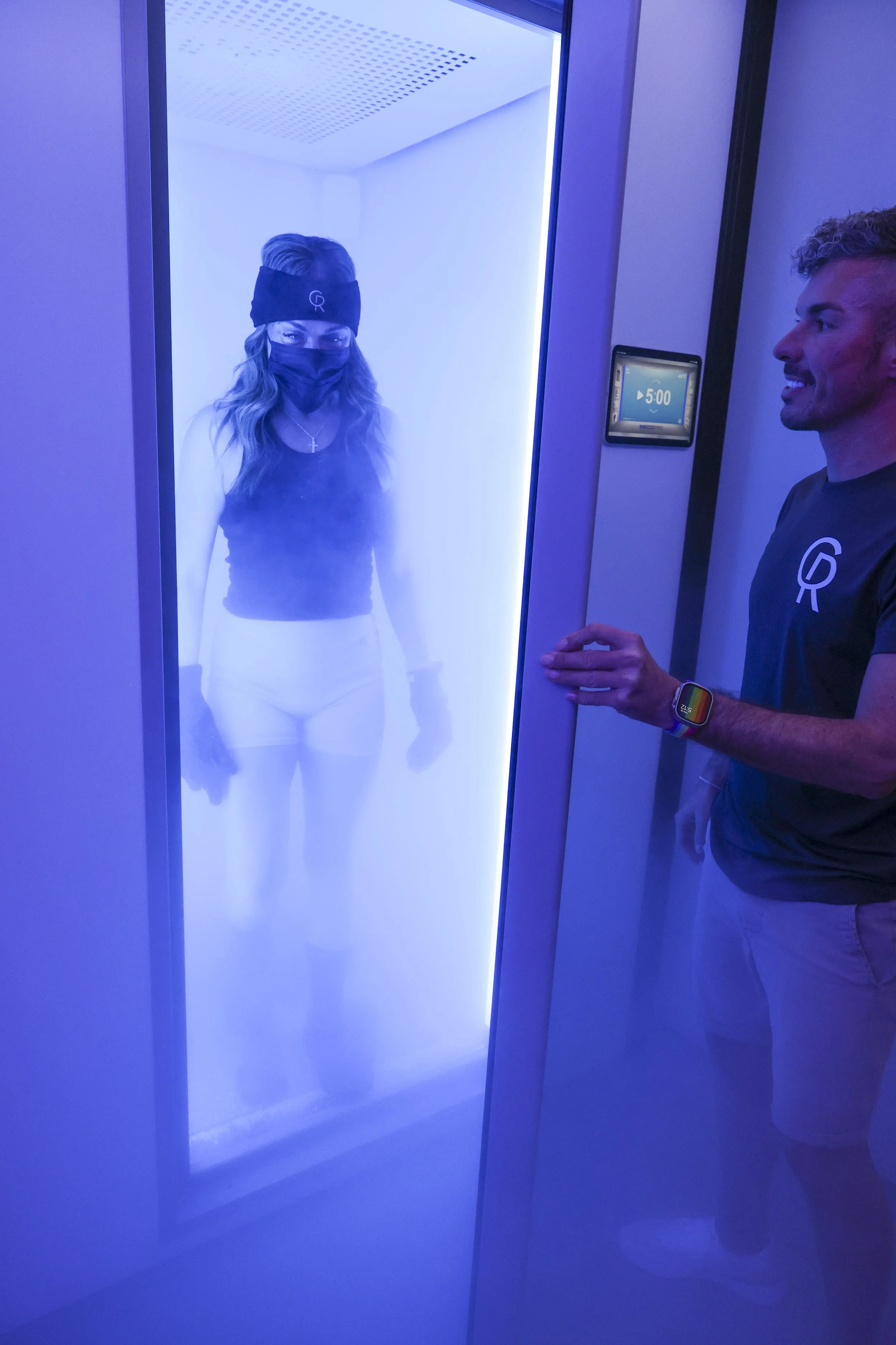 A client wearing protective equipment is standing inside The Cold Room's Mecotec full-body cryotherapy chamber at -87°C. Supervised by a staff member.
