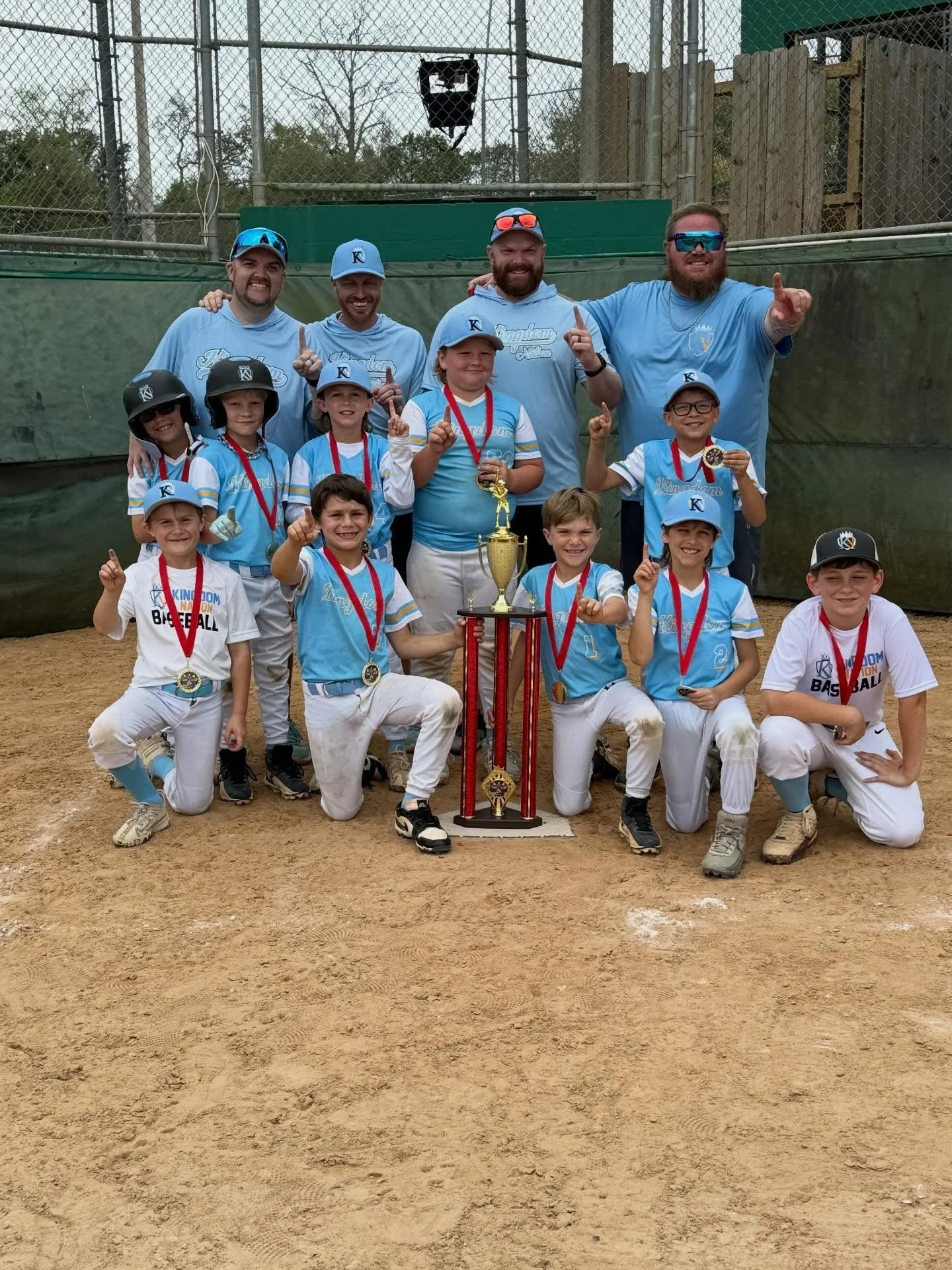 A youth baseball team, including children and adult coaches, posing on a baseball field with a large trophy and medals. The team is dressed in blue uniforms and some are wearing baseball helmets. The background shows a chain-link fence and trees.