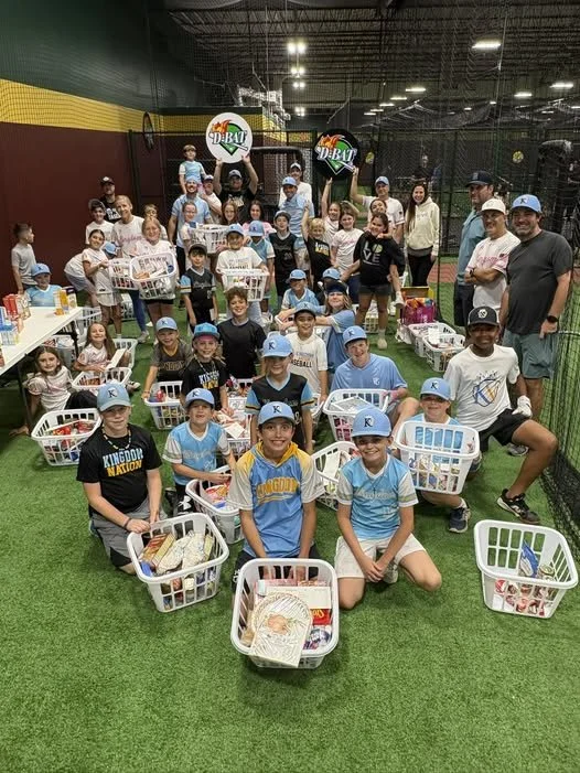 Group of children and adults at an indoor baseball event with baskets of supplies, some children wearing baseball jerseys and caps, background featuring nets and sports equipment.