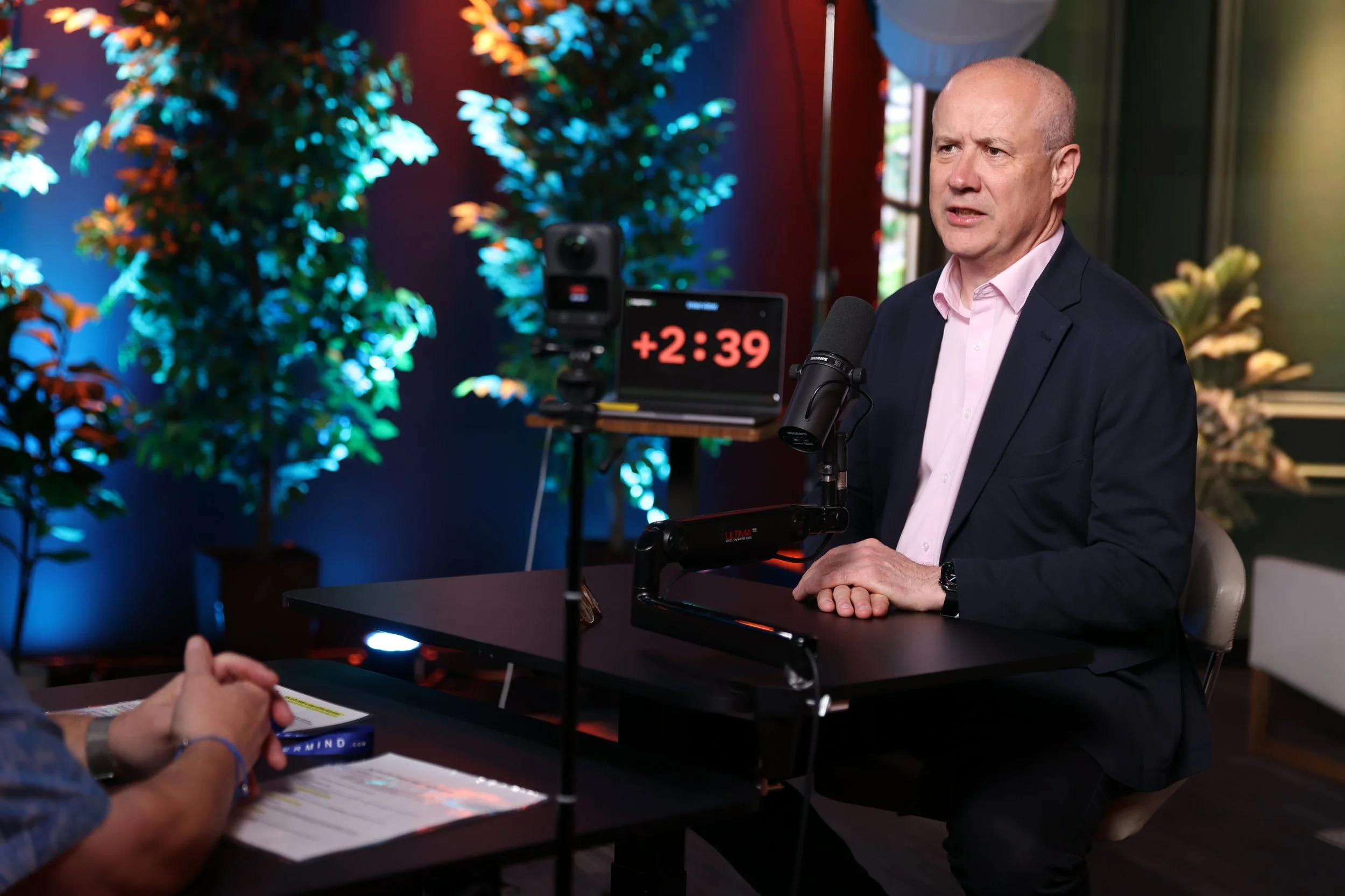 Stephen Mold in a suit and pink shirt speaking into a microphone during an interview or presentation.