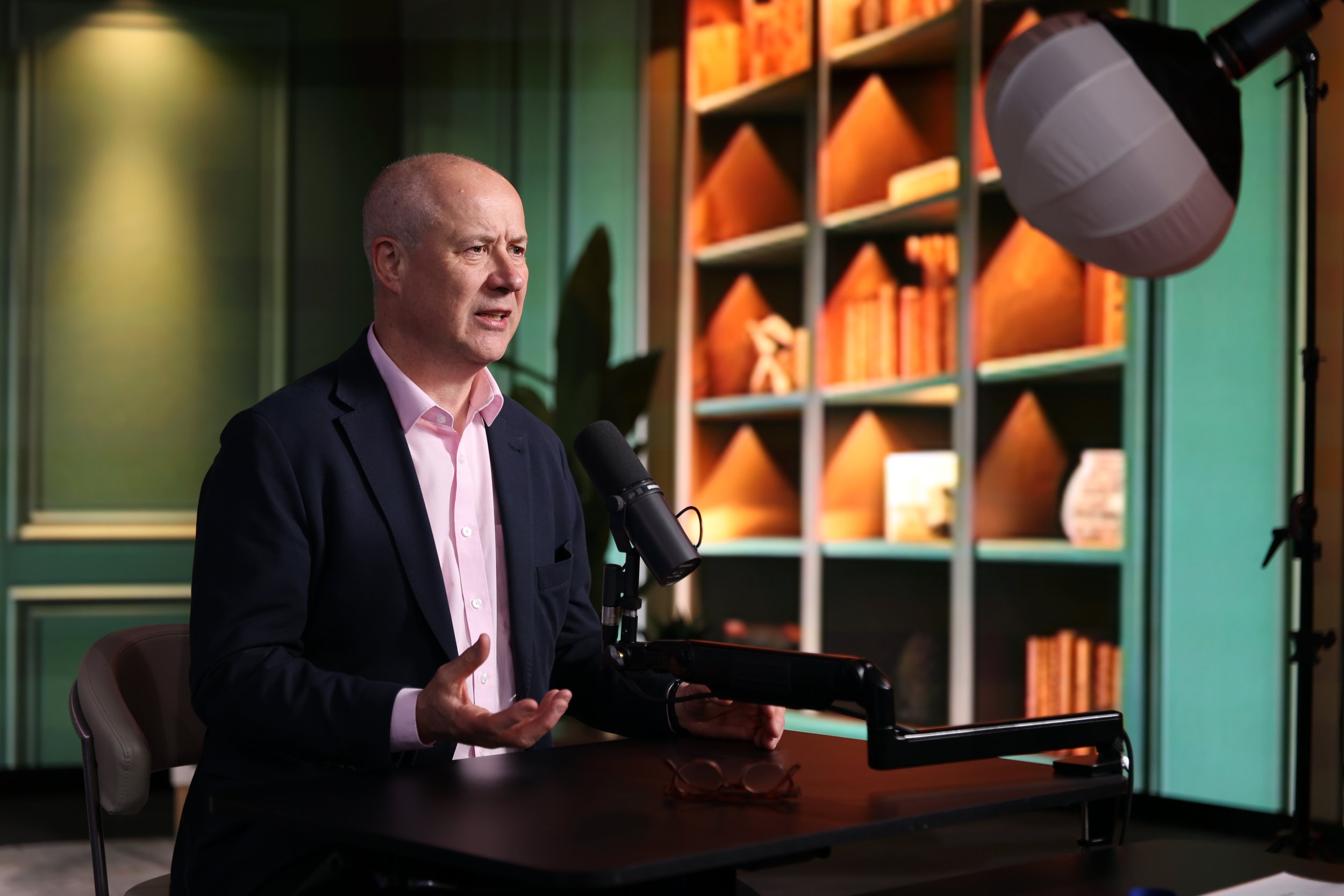 A man in a dark blazer and pink shirt speaking into a microphone during an interview or presentation, seated at a desk with a laptop and a pair of glasses, with a bookshelf and studio lighting in the background.