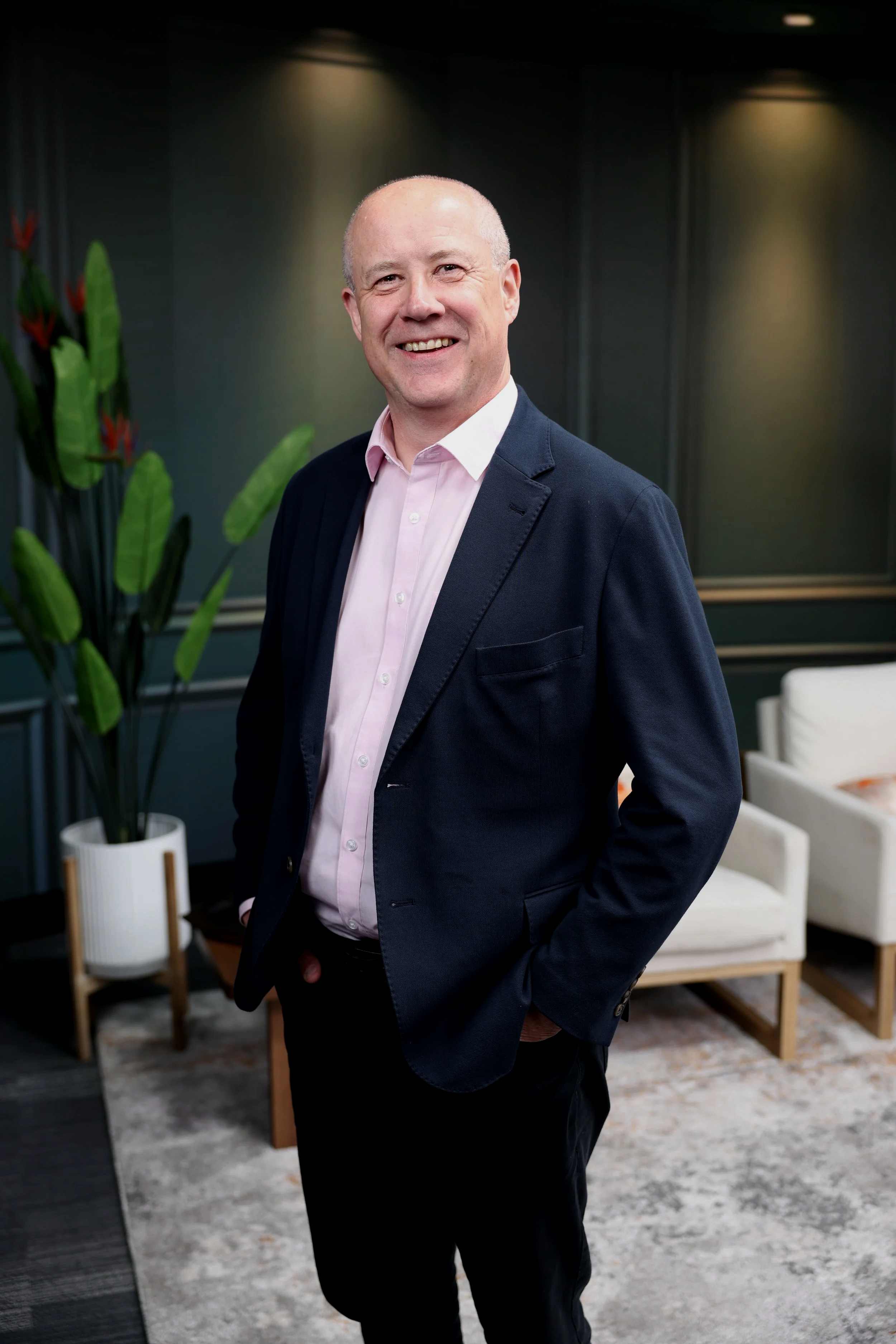 A smiling bald man wearing a light pink shirt and dark blazer standing in a modern office lounge with green plants, white chairs, and a rug.