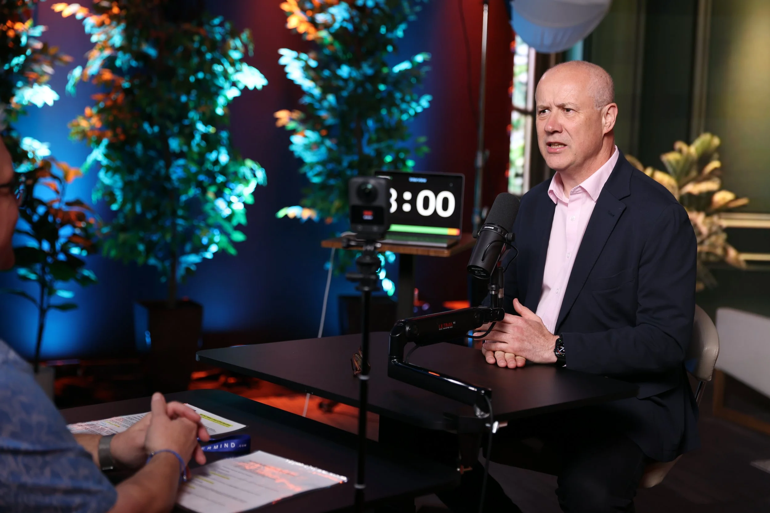 Stephen Mold in a suit and pink shirt speaking into a microphone during an interview or podcast recording.