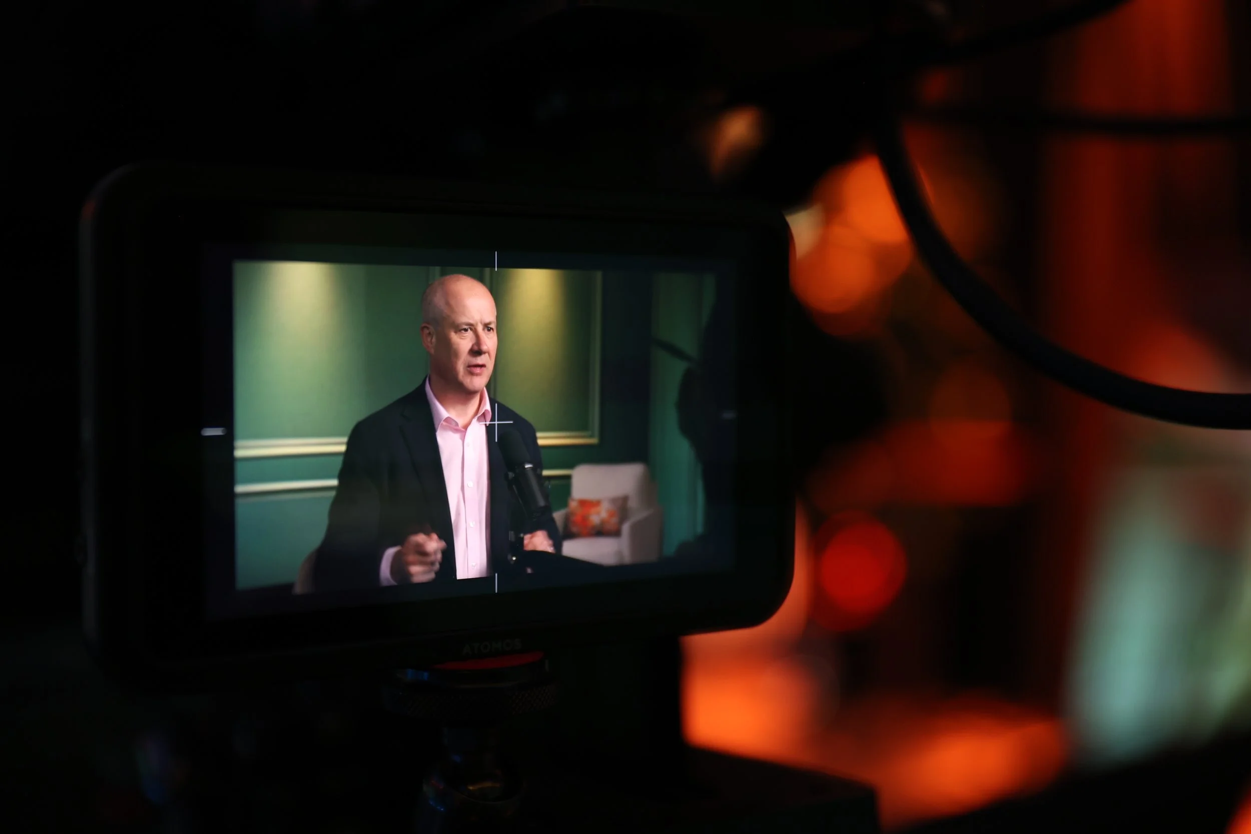 A camera screen shows a man in a suit speaking into a microphone in a room with green walls and a chalkboard.