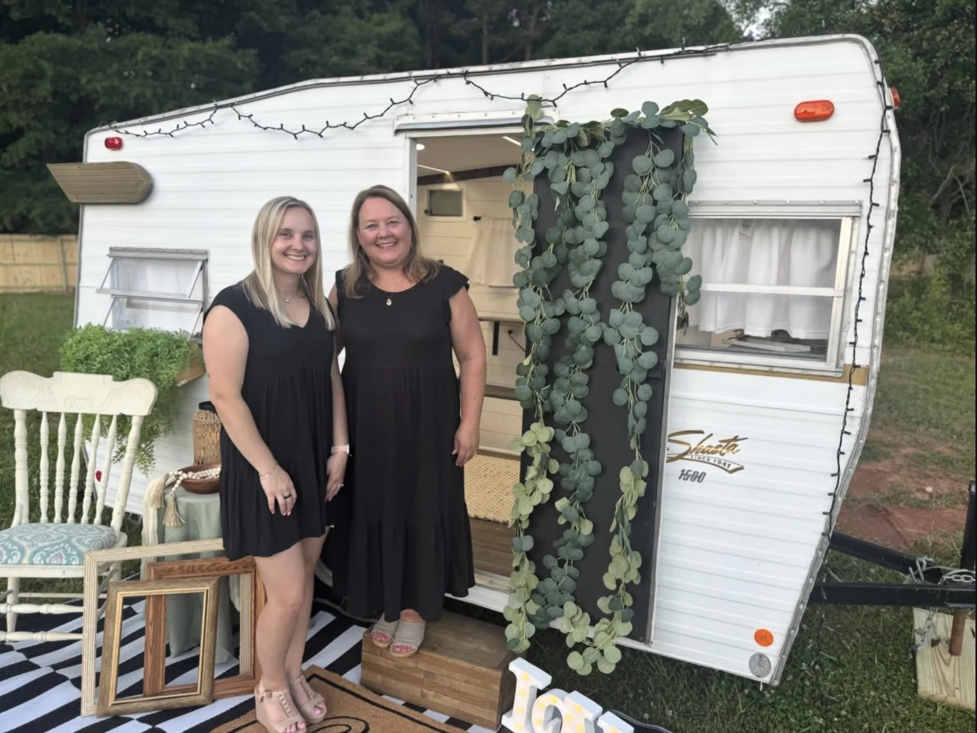 Two women dressed in black standing outside a white camper trailer decorated with hanging greenery. They are smiling and standing on a striped black and white rug, with a white chair and picture frames nearby. In the background, there are trees and a wooden fence.