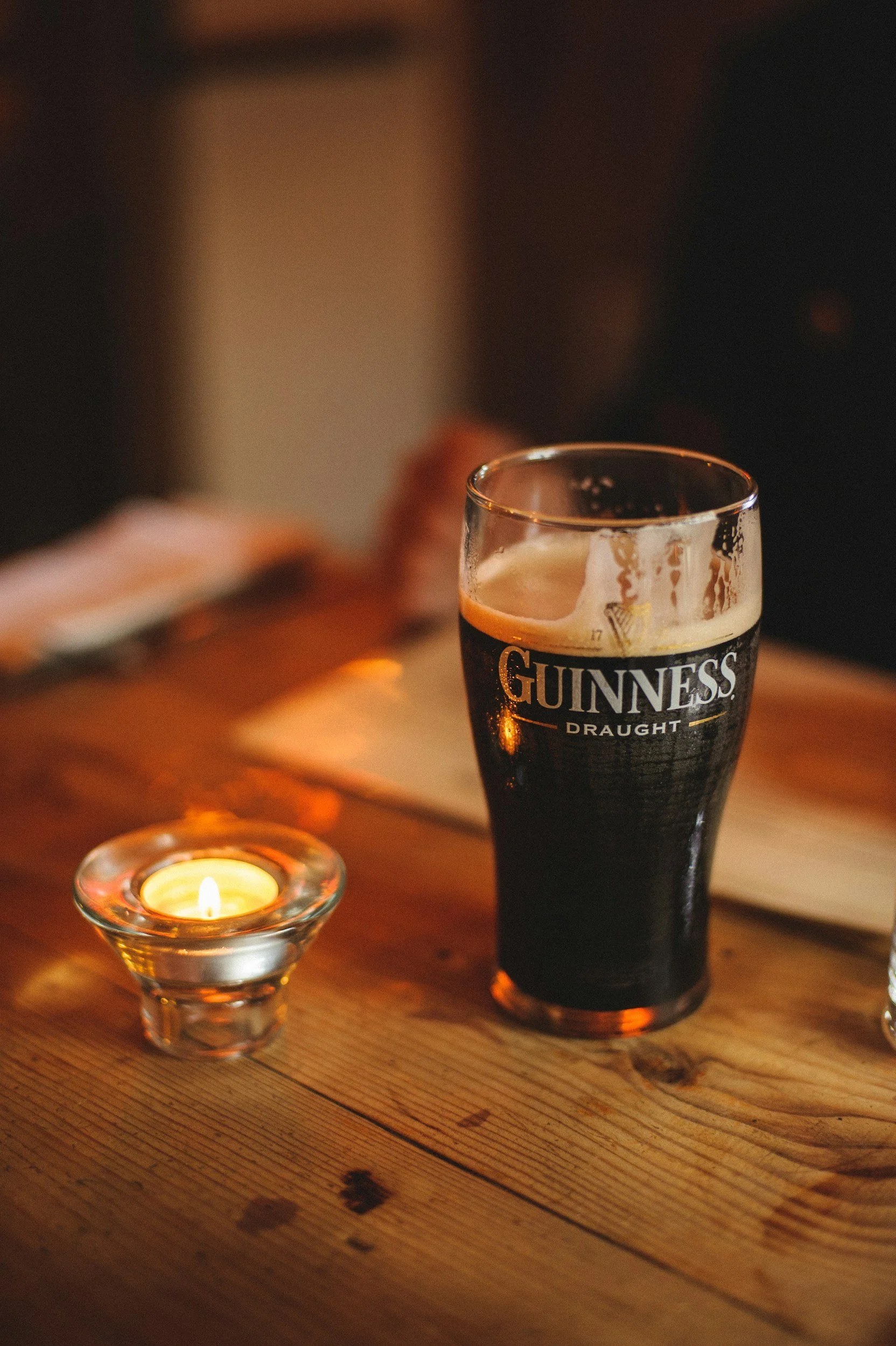 Ein Glas Guinness Bier auf einem Holztisch, daneben eine kleine brennende Kerze in einem Glas. Im Hintergrund ist die Umgebung dunkel, die Szene wirkt warm und gemütlich.