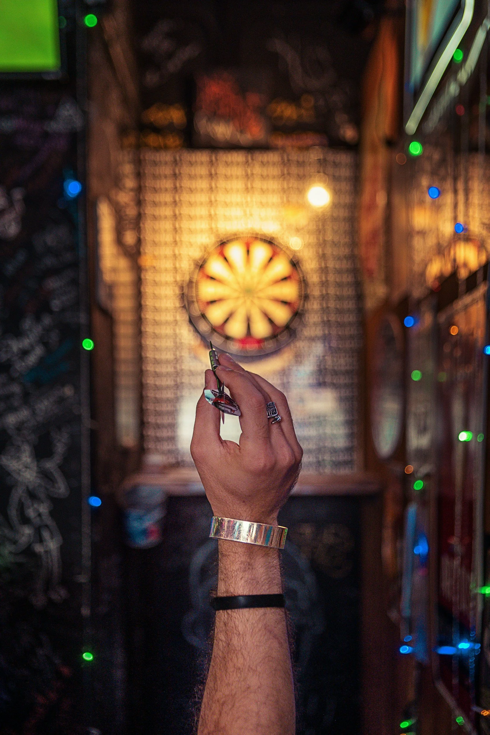 Nahaufnahme einer Hand, die eine Dartscheibe am Dartautomaten in einer Bar oder einem Spielraum hält. Im Hintergrund bunte Lichter und die Dartscheibe, die in der Nähe des Höchstpunktbereichs ist.