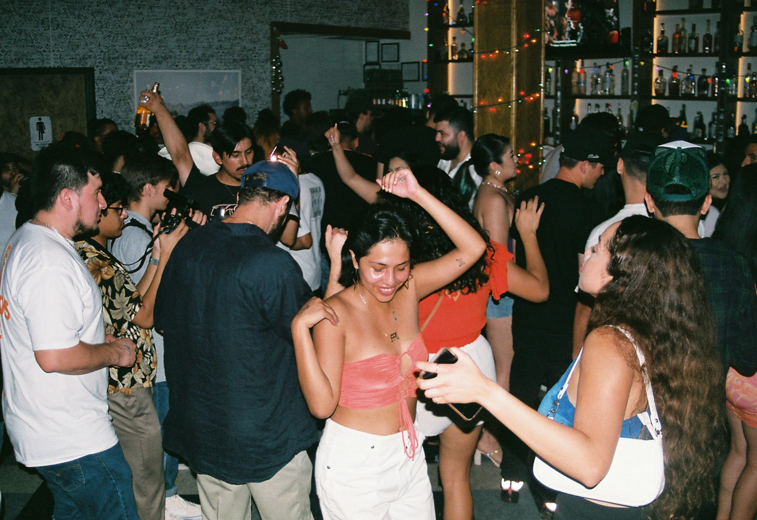 Menschen tanzen und feiern auf einer Party im Innenraum, mit bunten Lichtern und einem Barhintergrund.