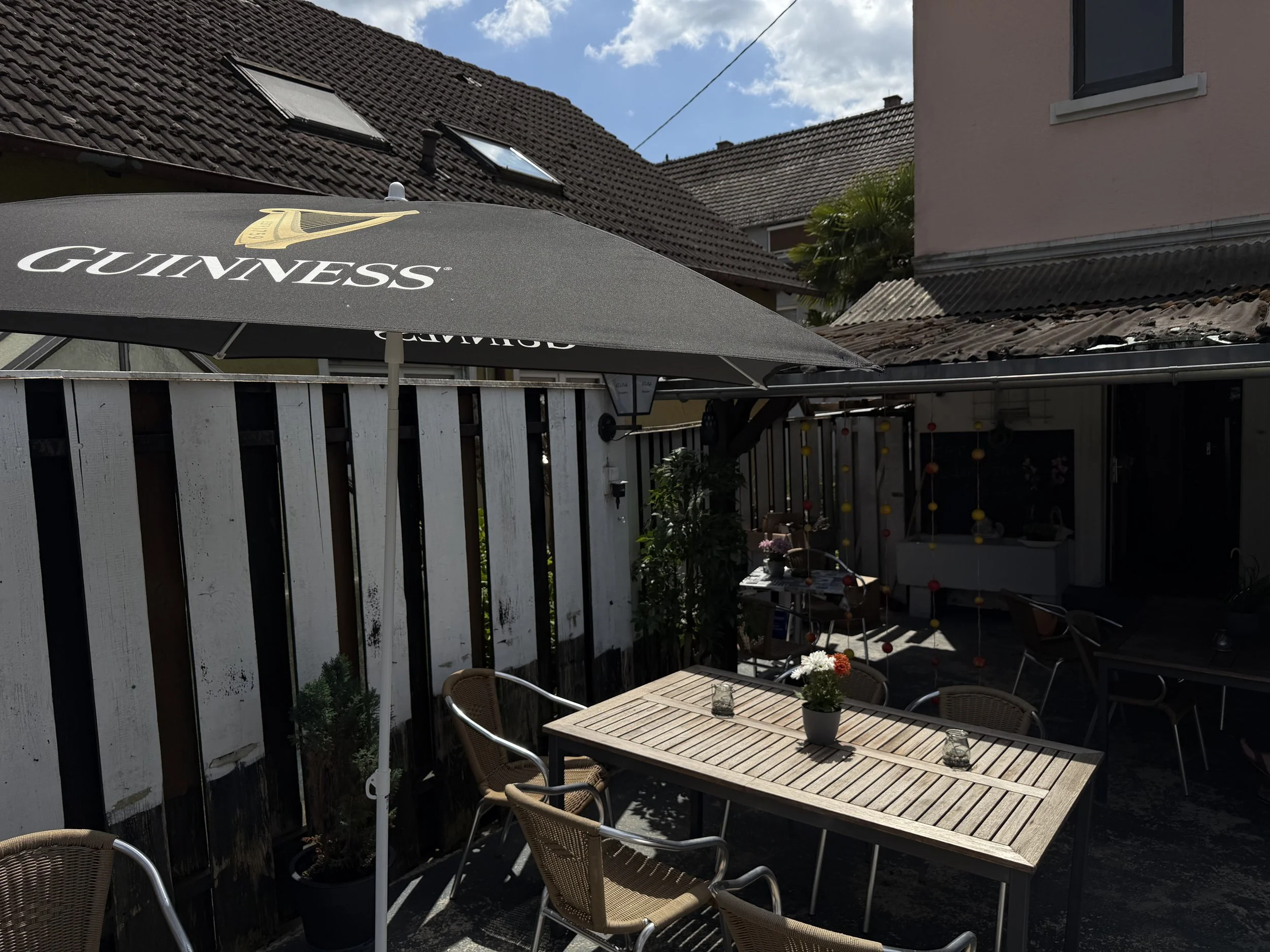 Ein Garten mit einem Holztisch, Stühlen, mehreren Pflanzen und einem Sonnenschirm mit Guinness-Logo, im Hintergrund Dächer und Himmel.
