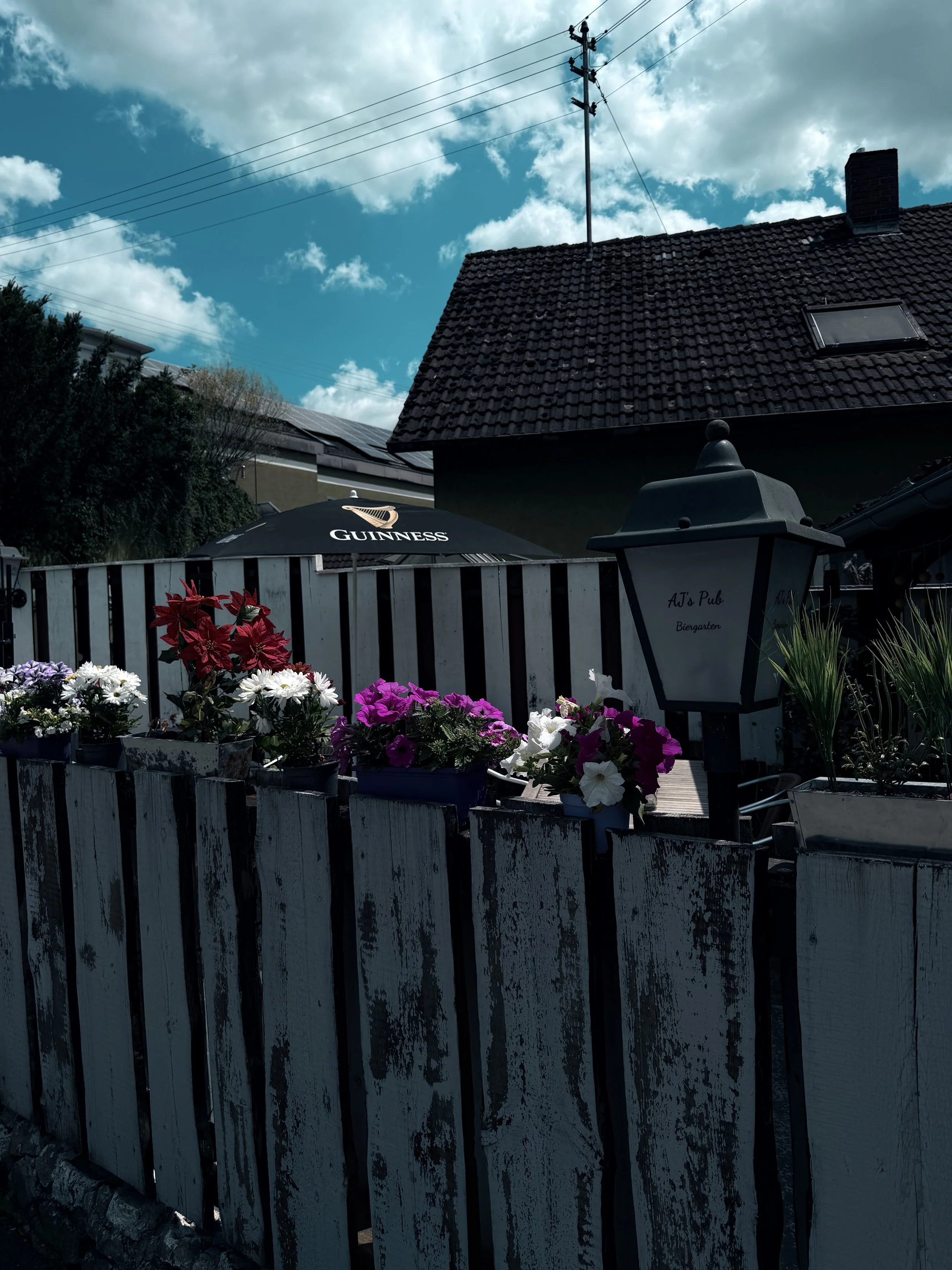 Garten mit bunten Blumen, schwarzer Sonnenschirm mit Guinness Logo, Laterne, Haus mit Dachfenster im Hintergrund, blaulichter Himmel mit Wolken.