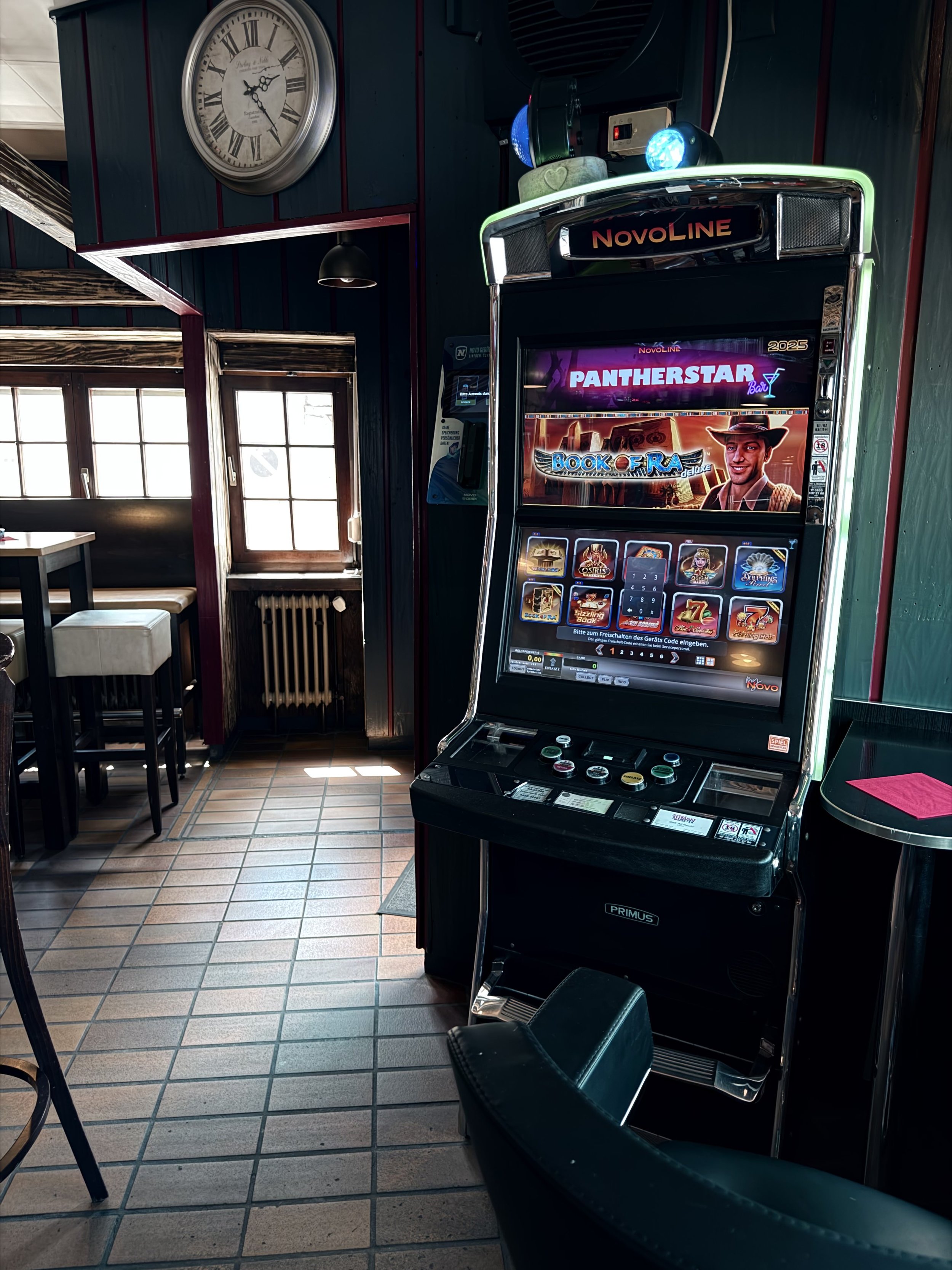 Innenraum eines Restaurants mit einem Spielautomaten, einem Wand-Uhr und Tischen mit Stühlen.