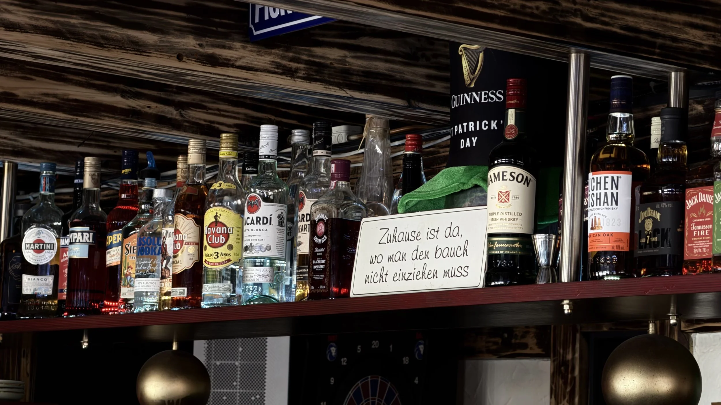 Blick auf eine Bar mit verschiedenen Spirituosenflaschen, darunter bekannte Marken wie Guinness, Jameson, Jack Daniel's, Jim Beam und andere. Es gibt eine Tafel mit deutschem Text und eine schwarz-weiße Irish-Flagge.