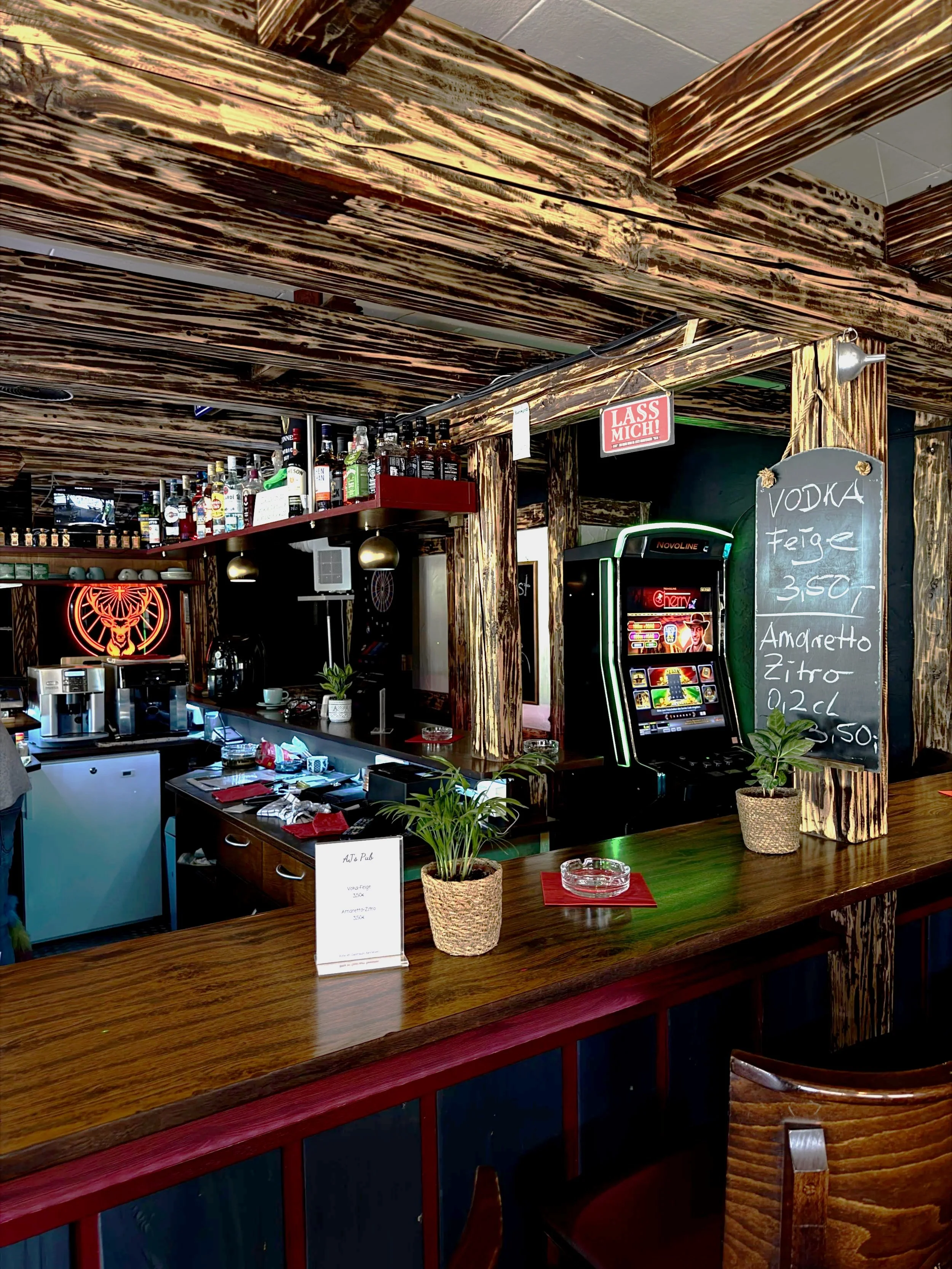 Blick auf eine Bar mit einem Holztheke, im Hintergrund: Flaschen mit Alkohol auf einem Regal, ein Spielautomaten und eine Menütafel mit Getränken. Es scheint ein Pub oder eine Bar zu sein.