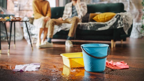 Two people sitting on a couch with cleaning supplies and water spilled on the floor in front of them.