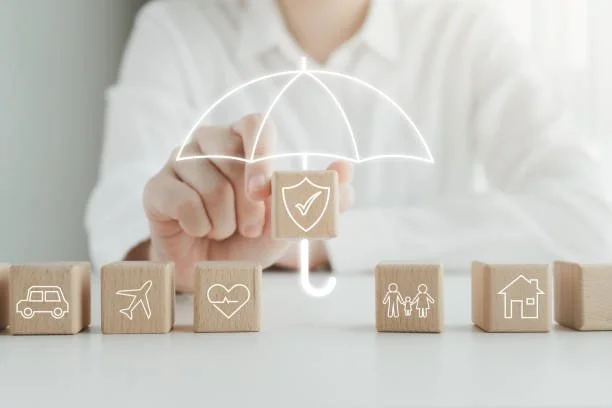 Person in white shirt placing a wooden block with a shield and check mark symbol under a digital umbrella icon, with other wooden blocks depicting vehicle, airplane, heart, family, and house symbols.