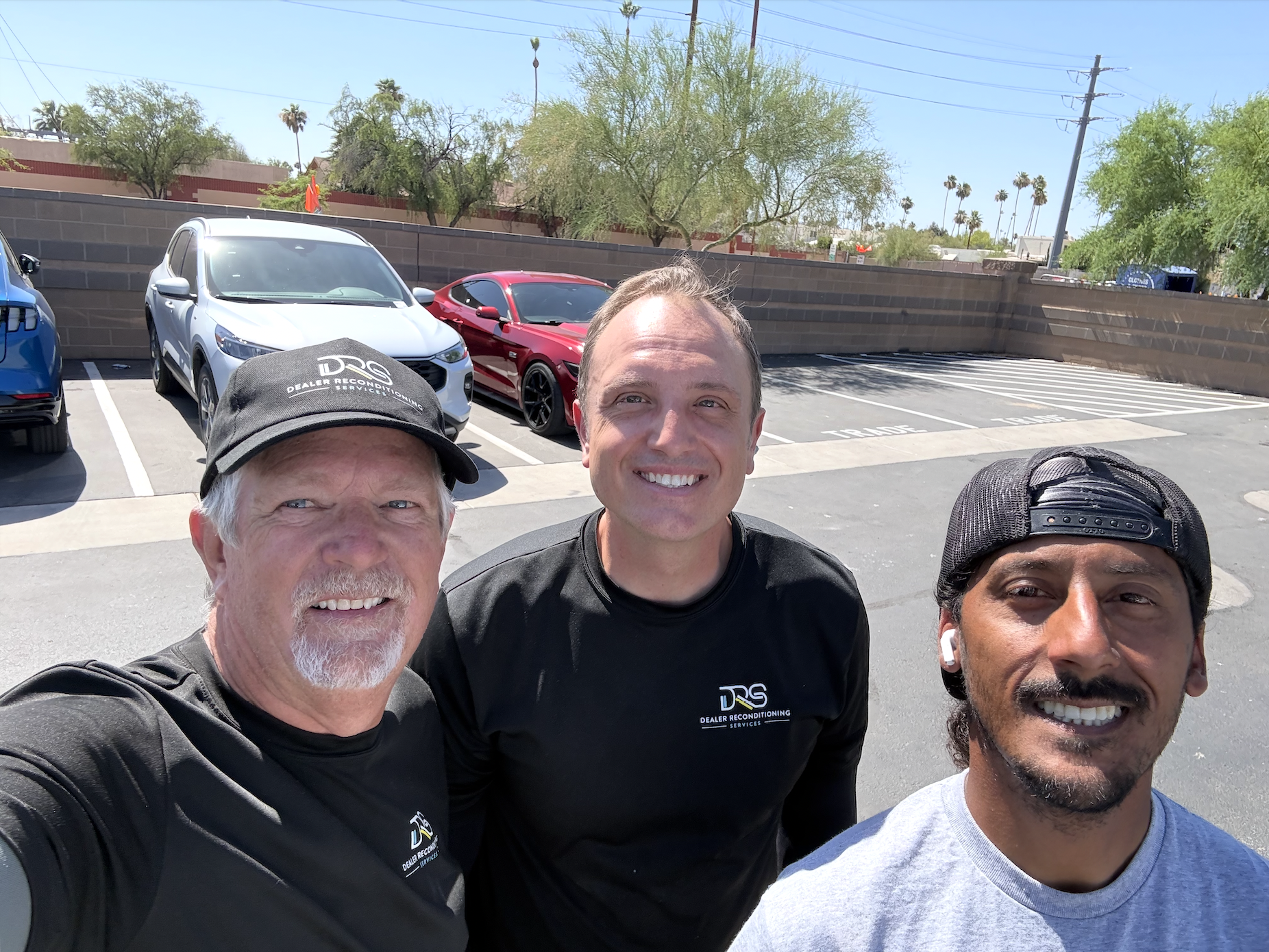 Three men standing outdoors, smiling at the camera, with a parking lot, colorful cars, trees, and power lines in the background.