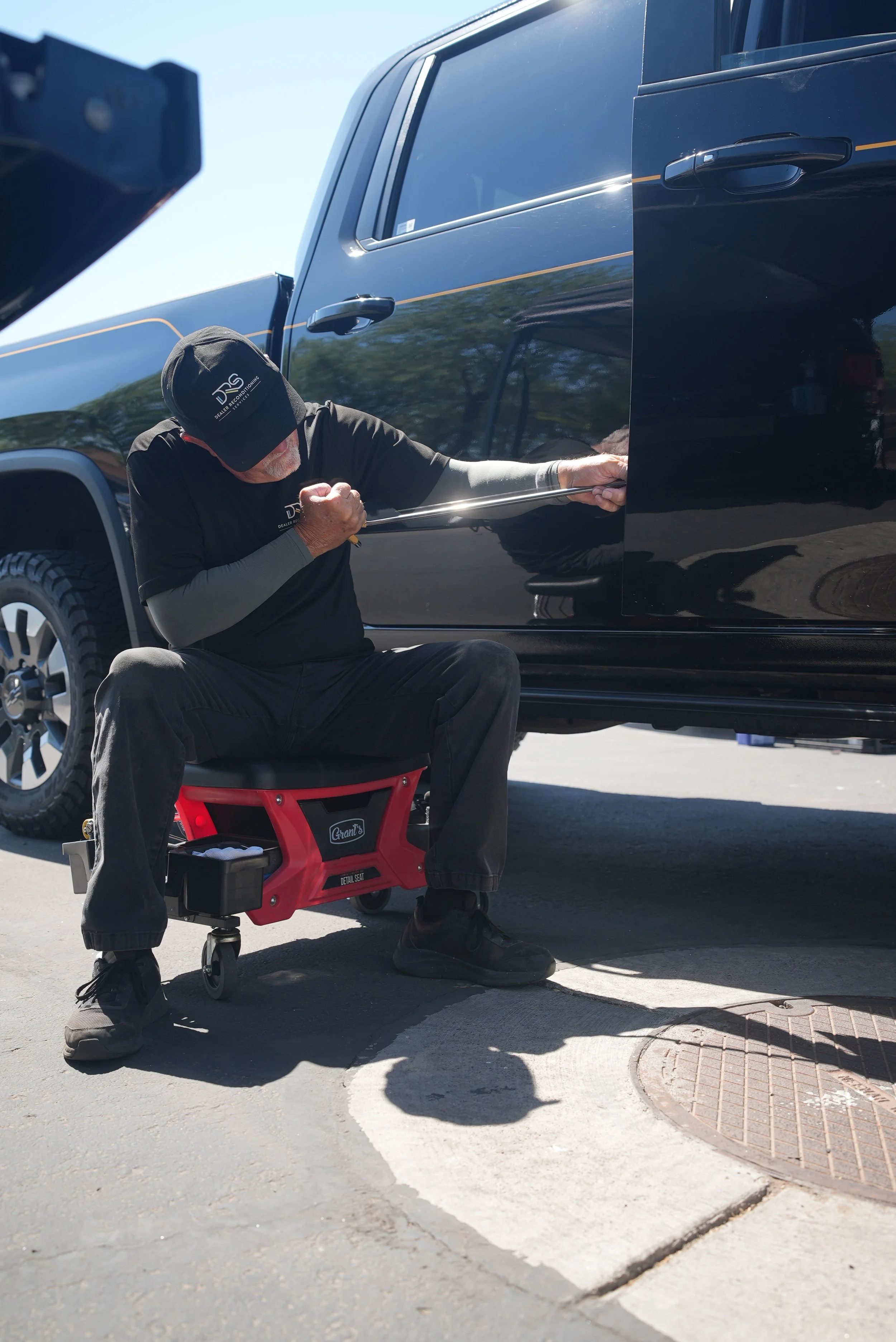 A man working PDR on a black pickup truck, seated on a red mechanic's stool. Paintless Dent Repair is getting the dents out of that truck in an efficient and nonintrusive manner