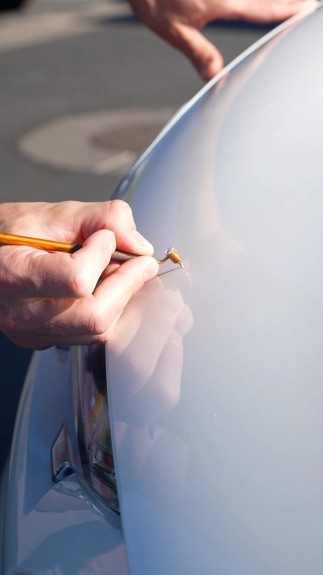 A person working on a silver car's body, using a small tool to carefully scrape or inspect the paint or surface.