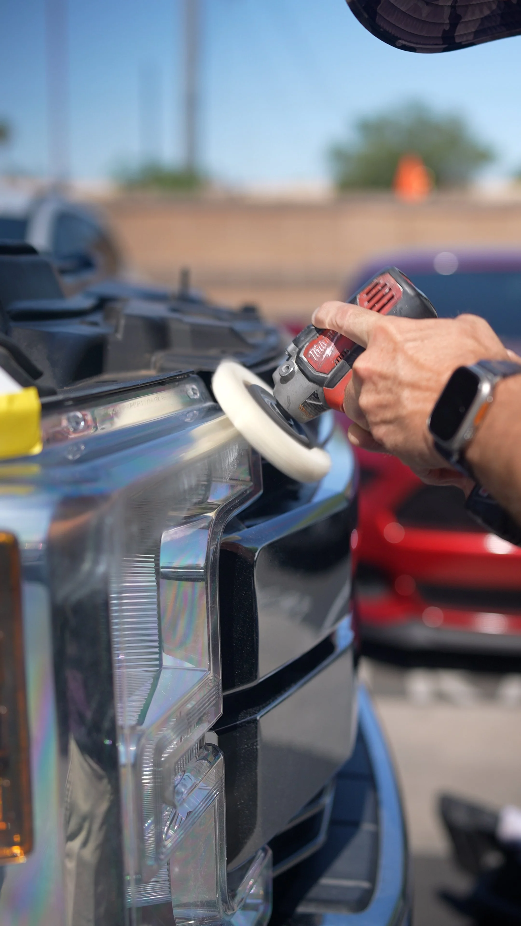 Close-up of a person's hand holding a cordless power drill, working on a vehicle's front panel at an outdoor location with a parking lot and cars in the background.