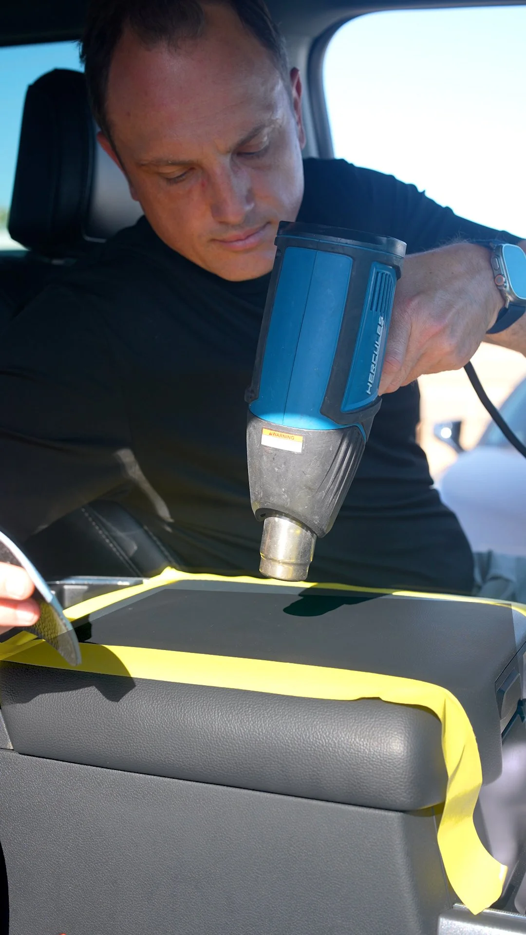 A man in a black shirt is using a heat gun to apply yellow tape to a surface inside a vehicle.