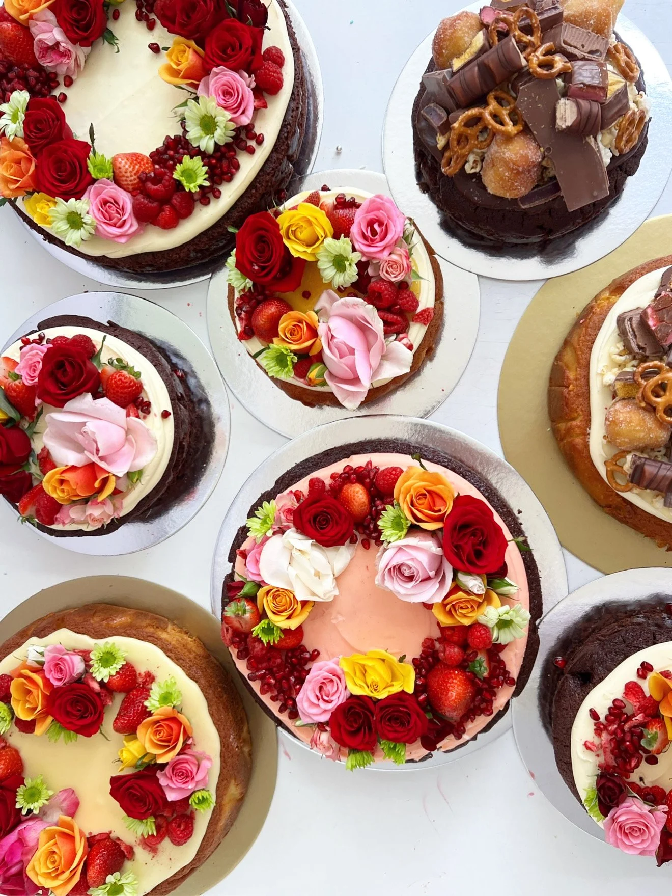 Various decorated cakes with flowers, berries, and chocolate toppings on a white surface.
