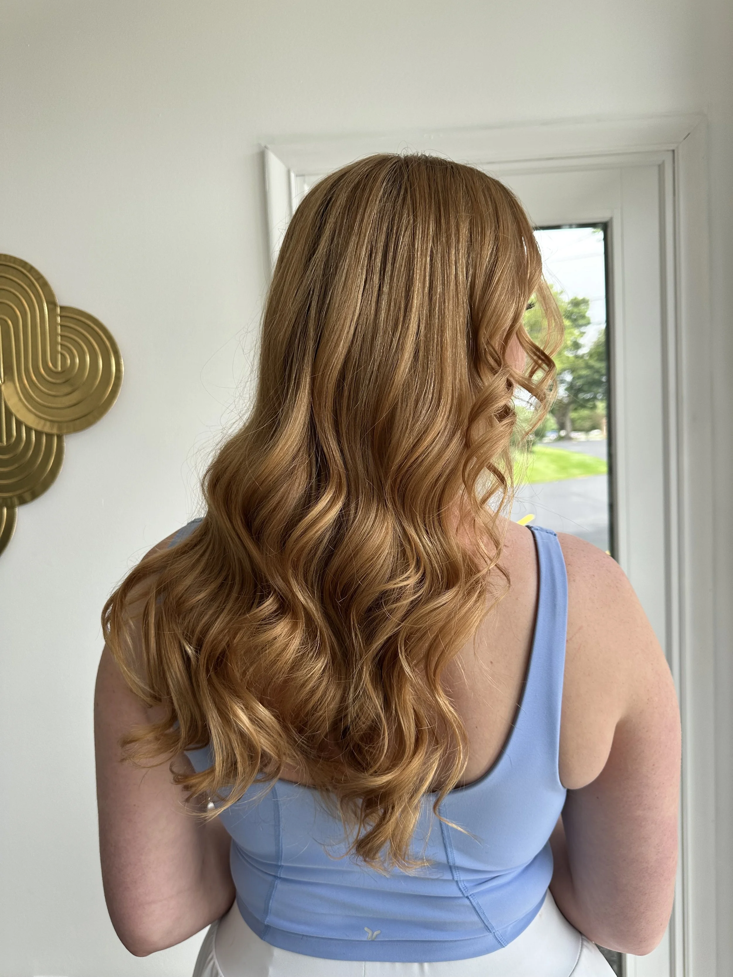 Back view of a woman with long, wavy, red hair, wearing a light blue tank top and white pants, standing in front of a white wall and a window showing green trees outside.