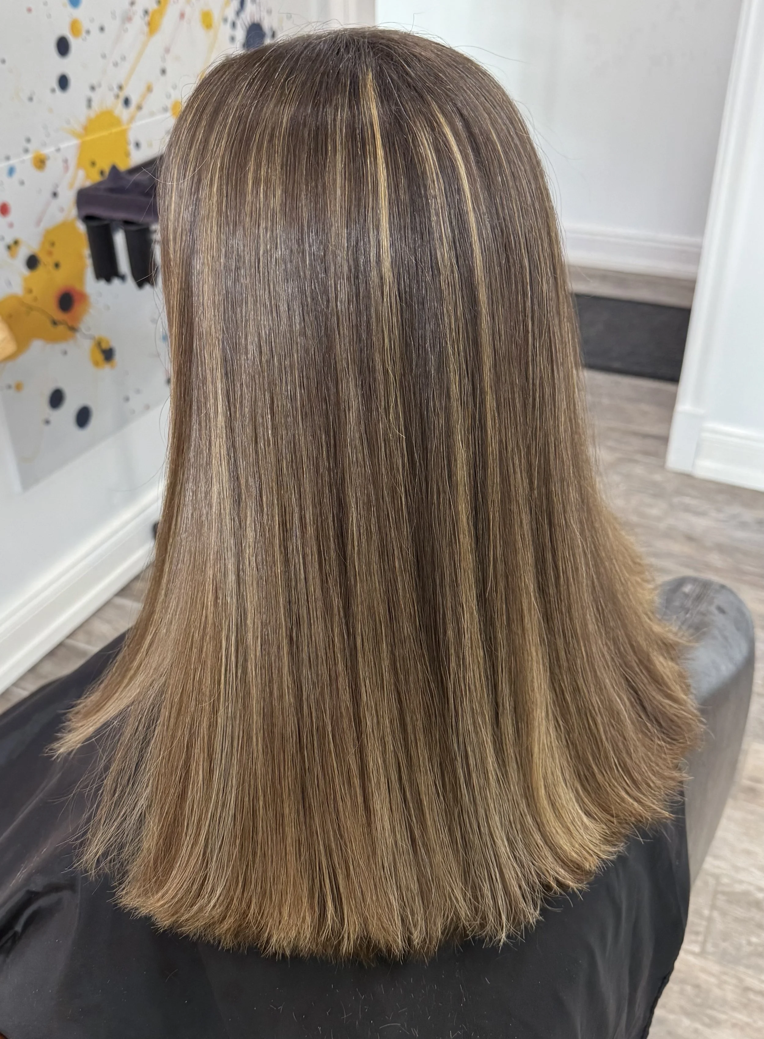 Back view of a person with long straight hair featuring blonde highlights and a brown base color, seated in a salon setting.