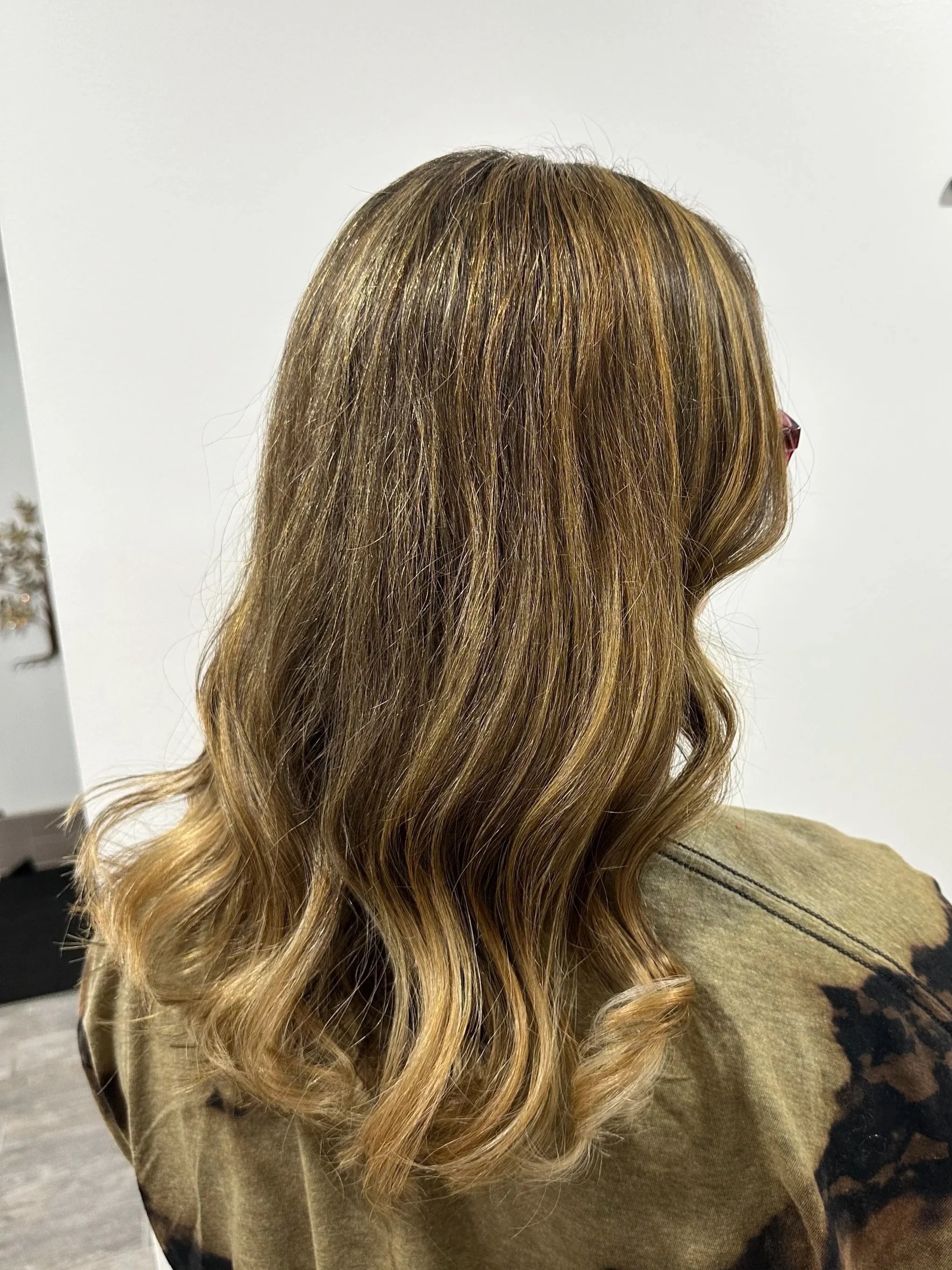 A woman with long, wavy hair that is light brown with blonde highlights, wearing a beige or olive green top with a floral or patterned black design on the sleeve.