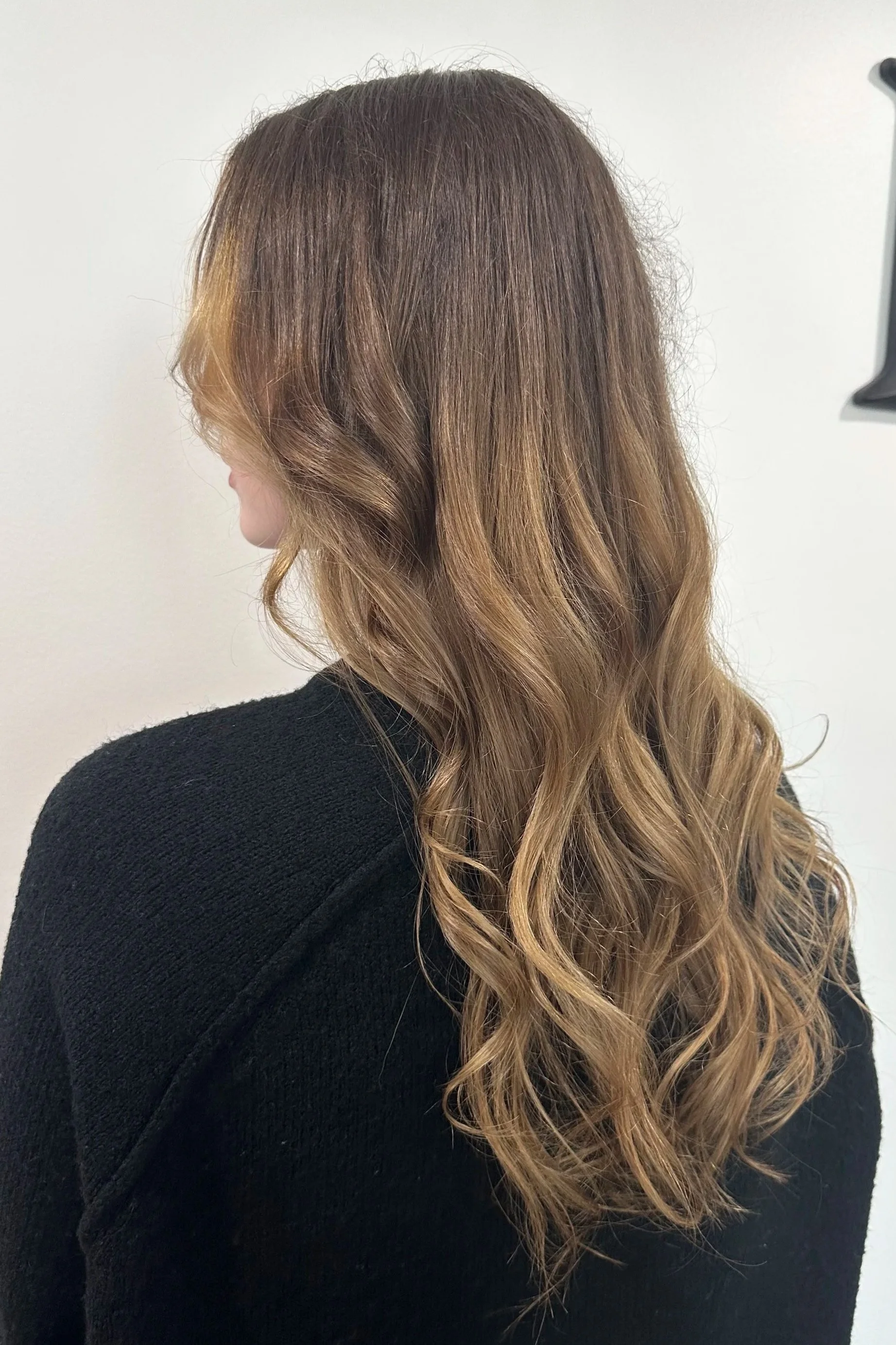 Back view of a woman with long, wavy, ombre hair transitioning from dark brown at the roots to blonde at the tips, wearing a black top.