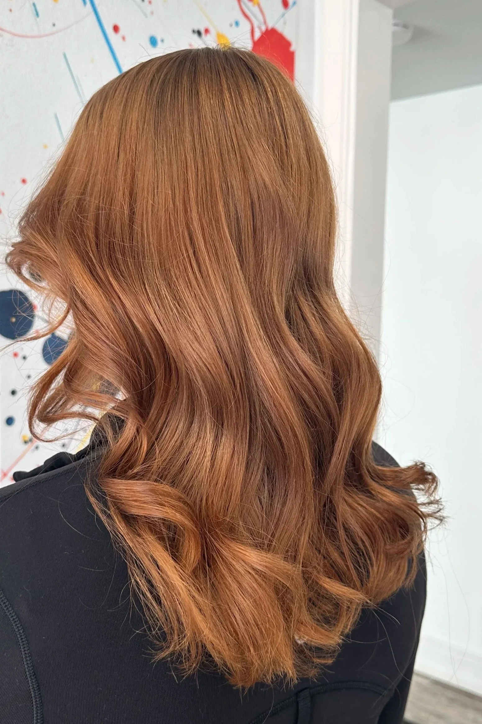 Wavy, shoulder-length red hair on a person wearing a black top, with a colorful, abstract background featuring splashes of red, black, and yellow paint.