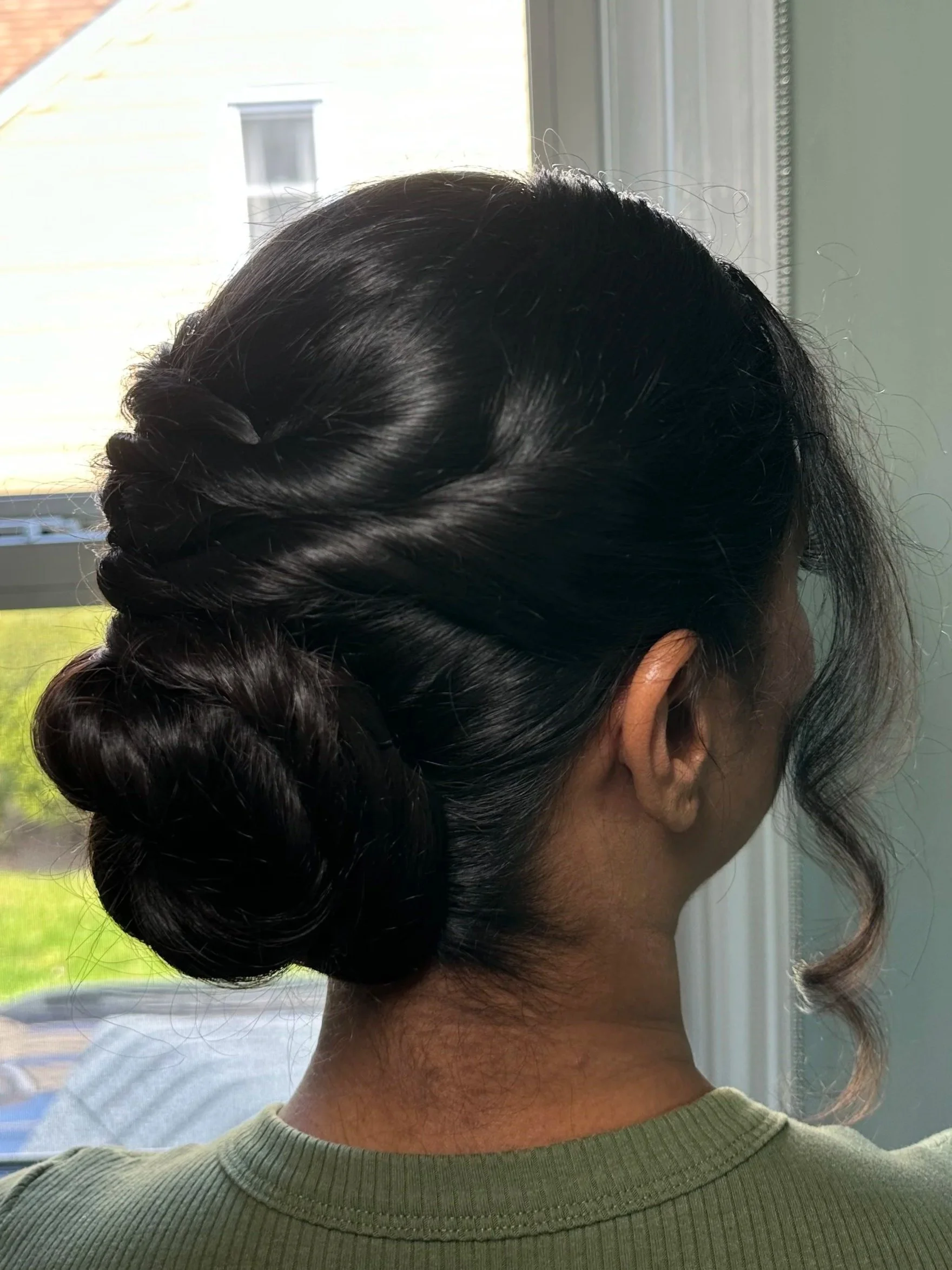 Close-up view of a woman's dark, shiny hair styled in an elegant twisted updo, with some loose strands around her face and tip of her ear visible, on a background with window blinds and a building outside.