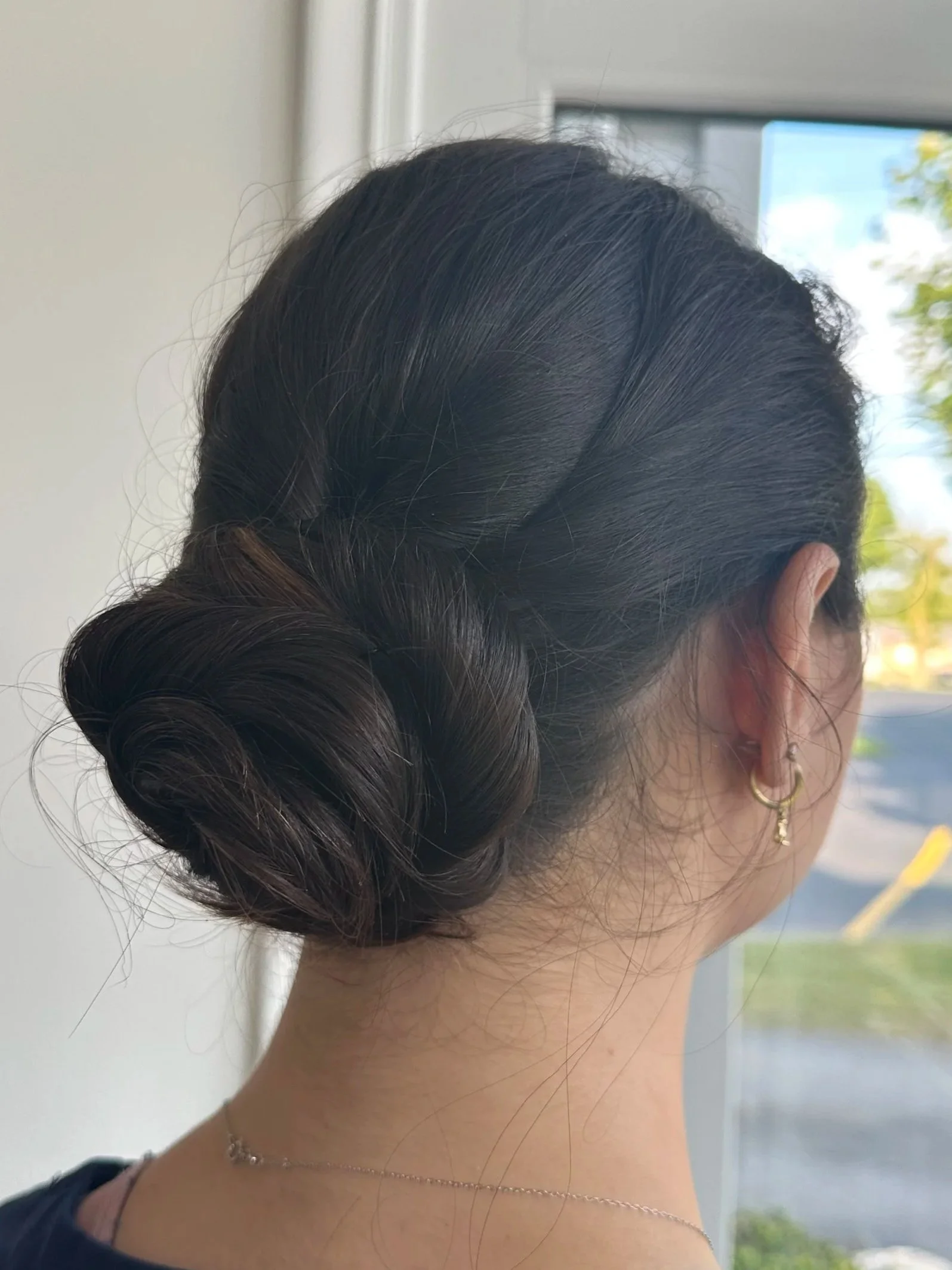 The image shows a hairstyle featuring vintage-inspired rolled-up hair at the back of the head, with some visible waves and shiny dark hair, worn by a woman with earrings and a necklace.