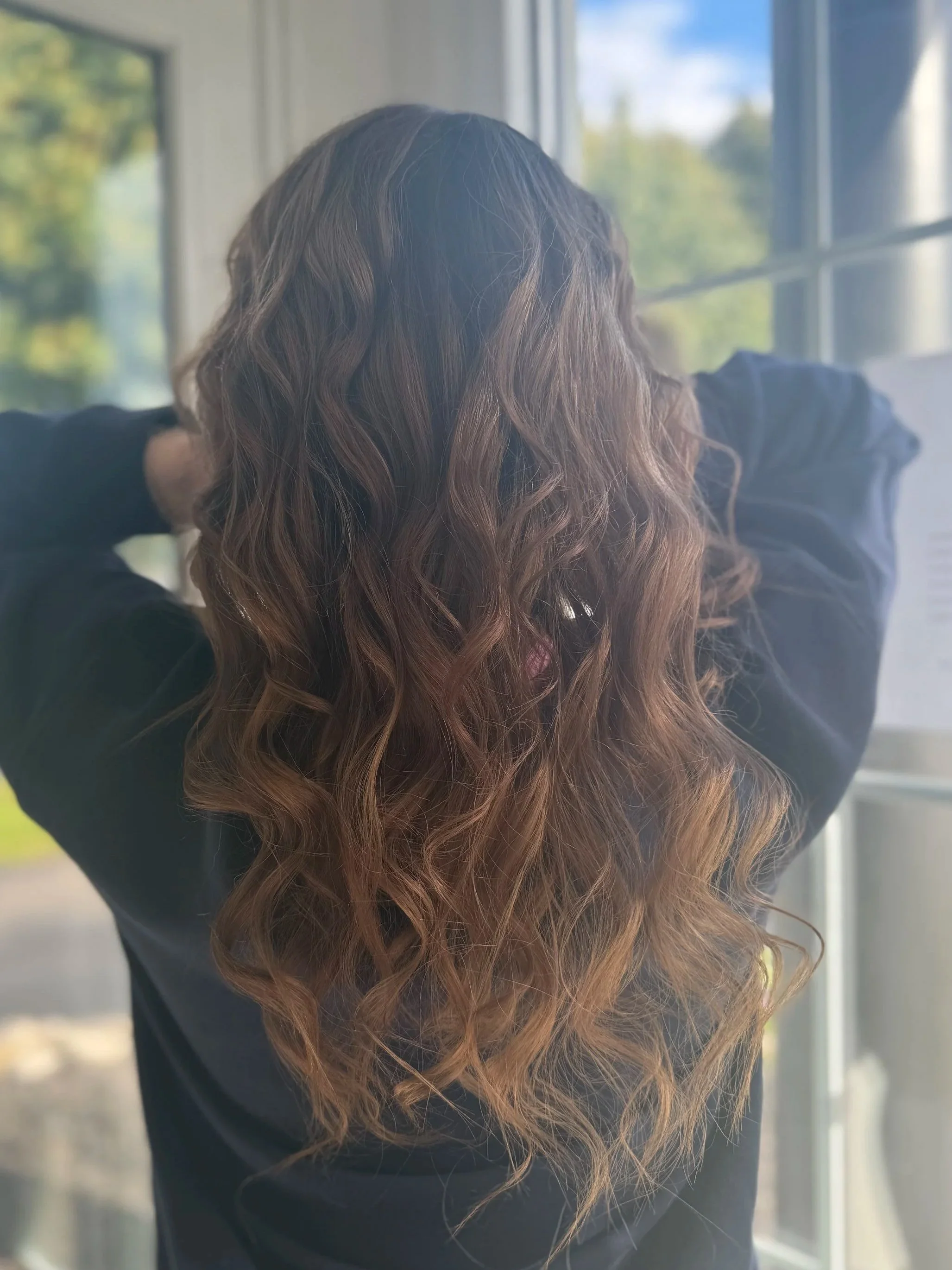 Back view of a woman with long, wavy, brown hair standing by a window, looking outside on a sunny day.