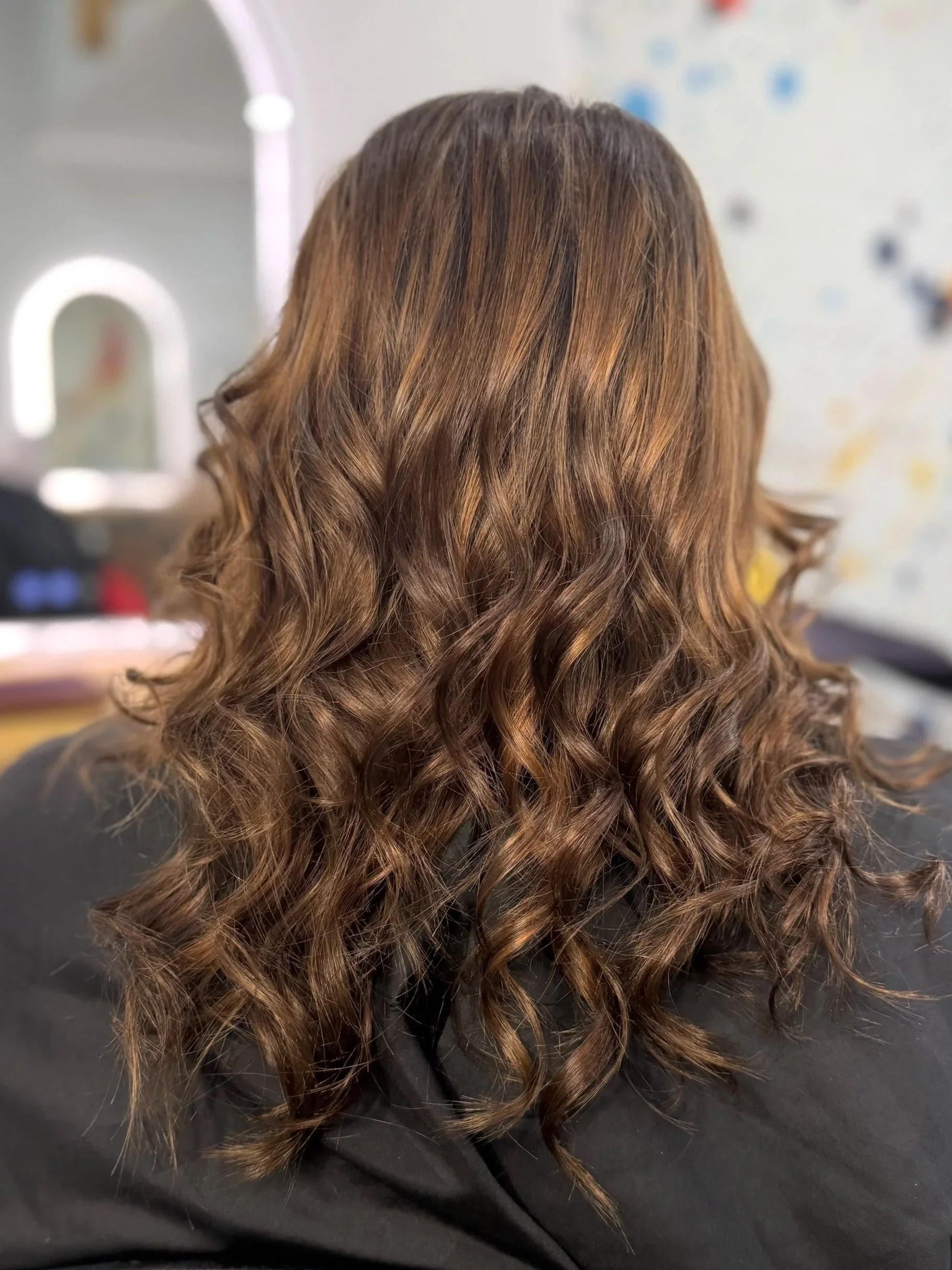 A person with long, curly, brown hair sitting in front of a mirror, possibly at a hair salon.