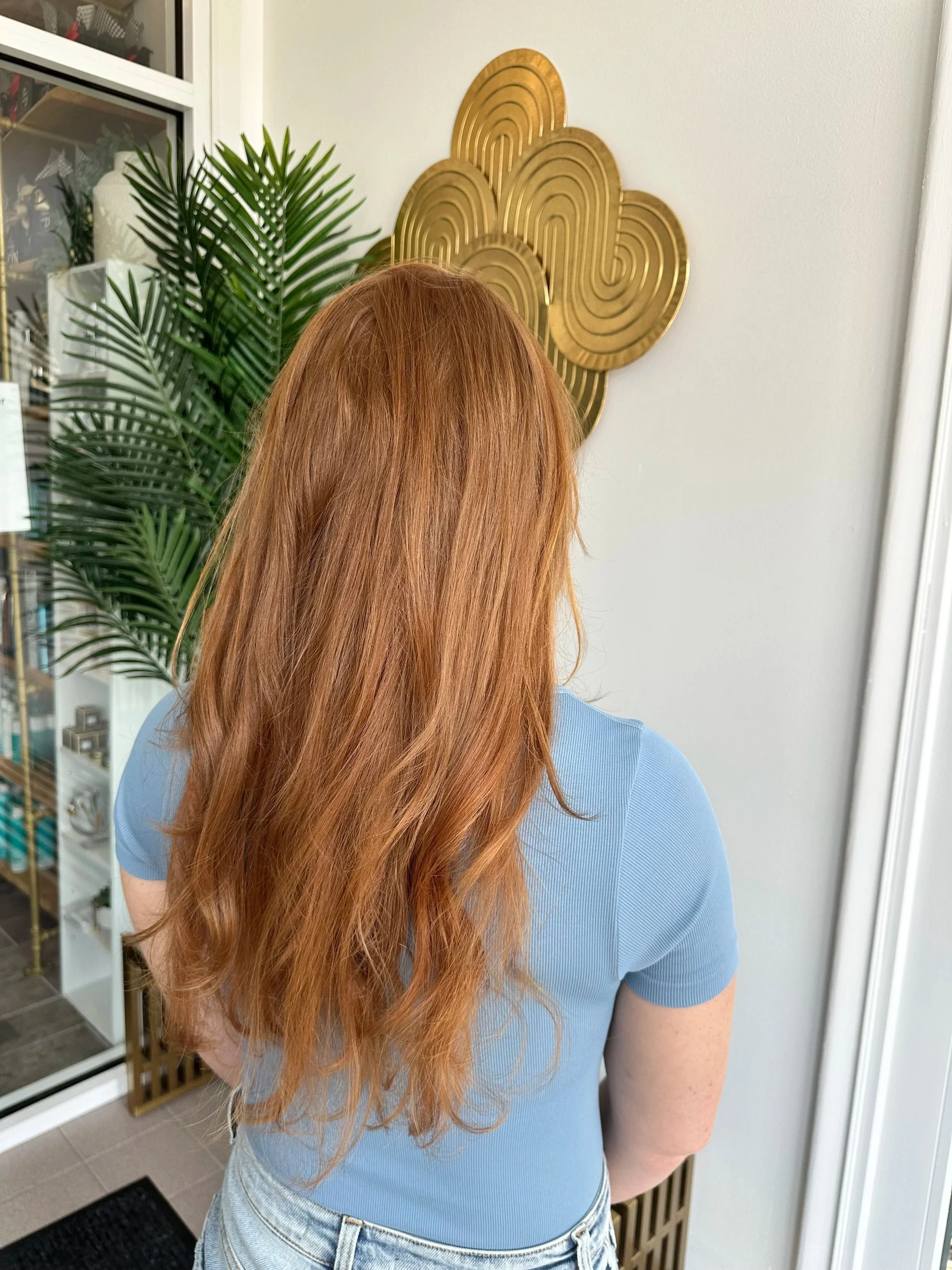 Back of a woman with long, wavy red hair, wearing a light blue shirt and denim jeans, standing indoors near a decorative gold wall art and a green potted plant.